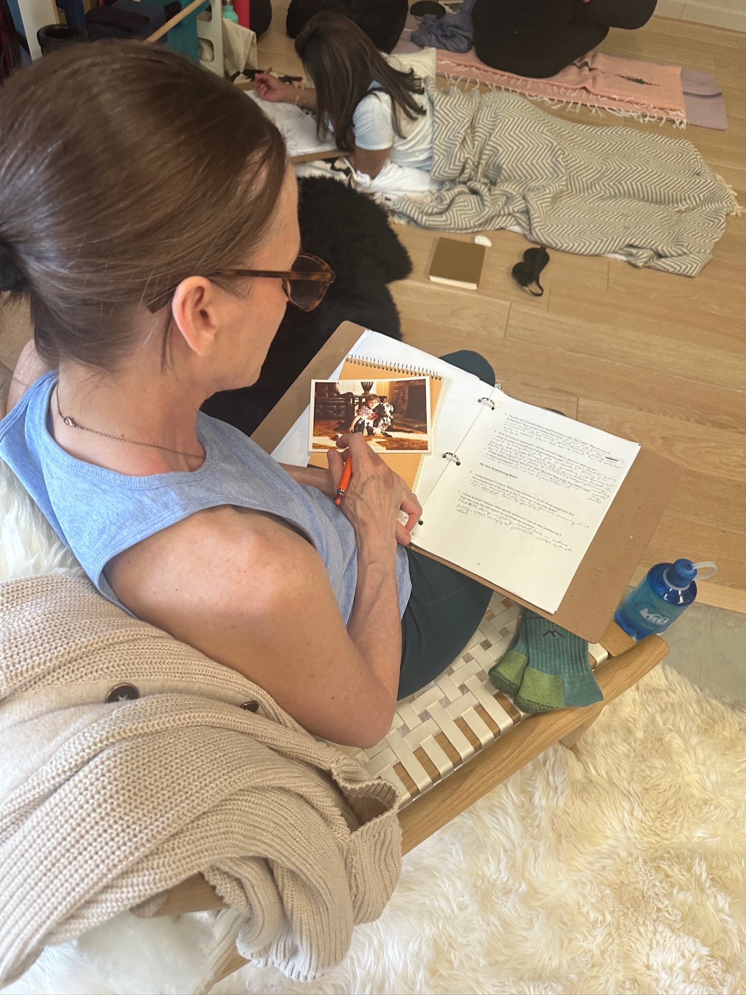 A woman with glasses and brown hair, wearing a beige sweater, is sitting on a cushioned wooden chair with a white furry rug, looking at a scrapbook or photo album with a childhood picture of a boy with dogs. Another woman is lying on the floor in the