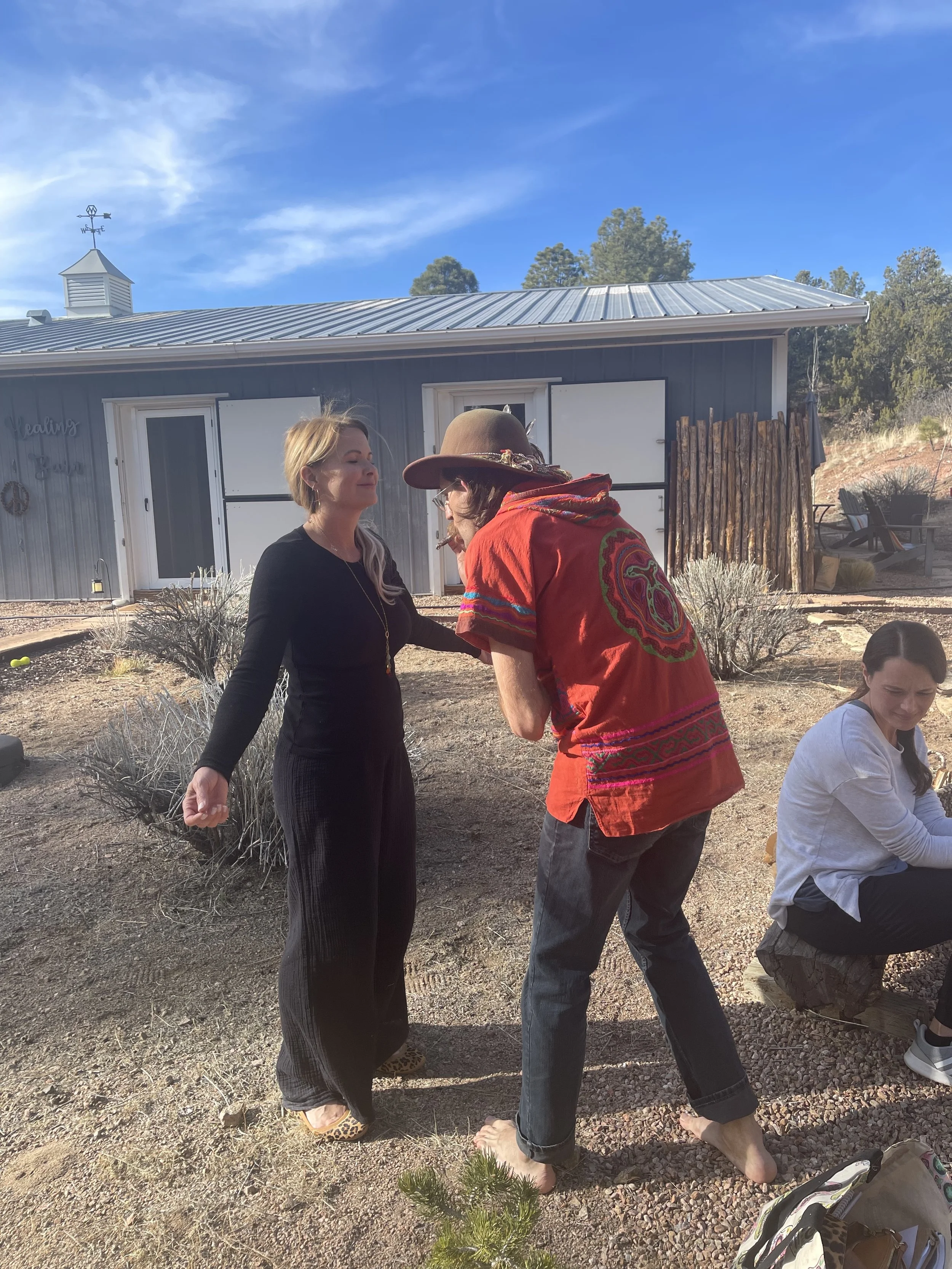 Two women engage in a conversation outdoors beside a house with a metal roof, while a third woman sits with her back on the ground nearby. The setting appears to be a backyard with trees and outdoor furniture in the background.
