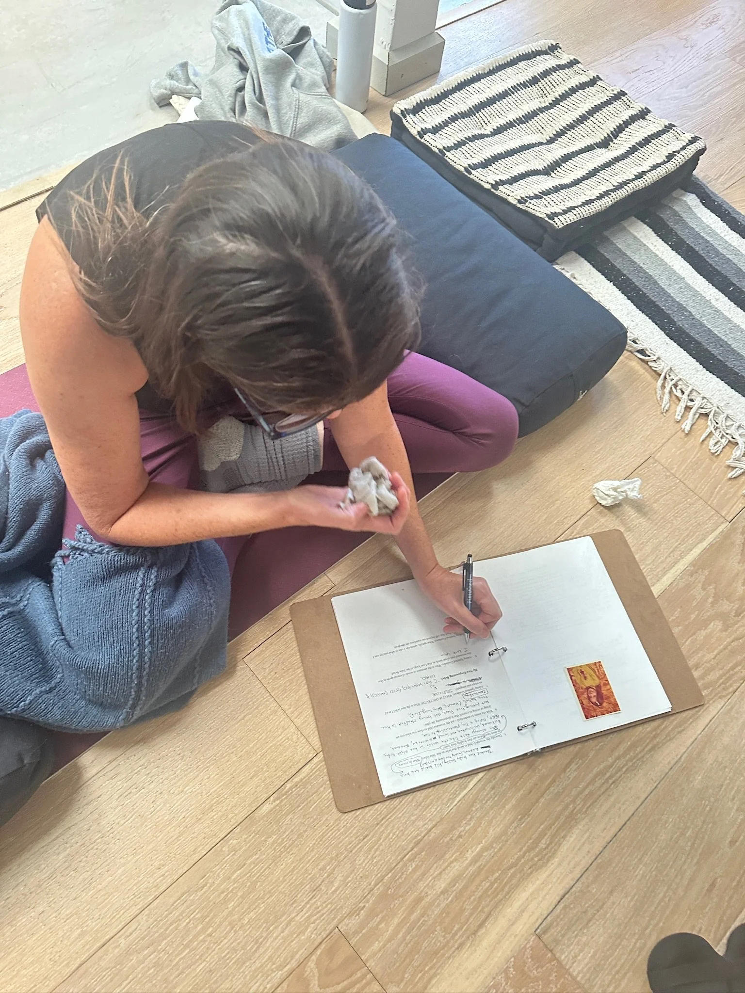 A person sitting on the floor, writing in a binder with handwritten notes, surrounded by crumpled paper and blankets, near a black and white striped cushion and a black mat, on a light wooden floor.