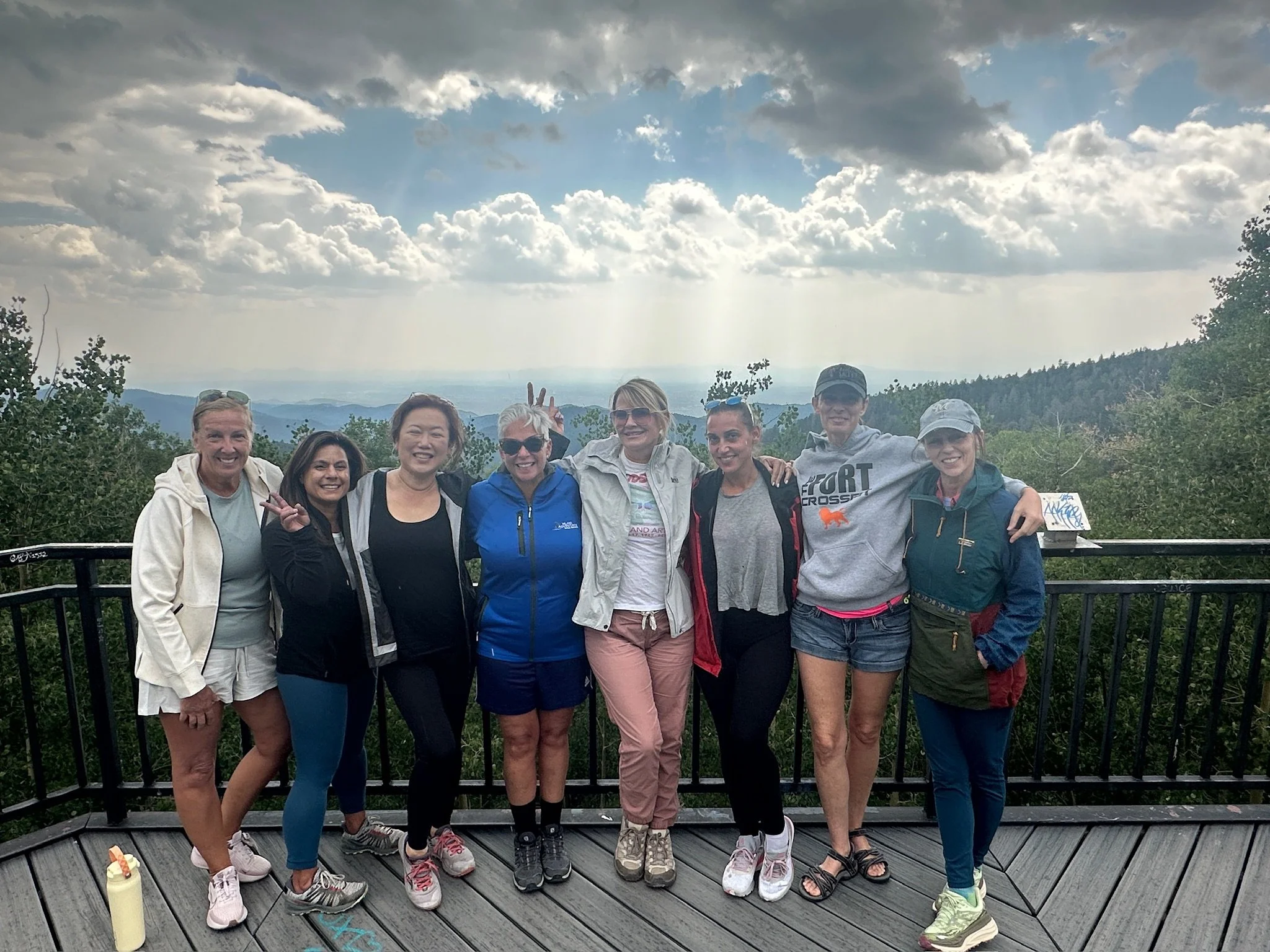 Group of nine people, mostly women, standing on a wooden viewing platform outdoors with mountains and a cloudy sky in the background. They are smiling and posing for the photo, dressed in casual outdoor clothing.