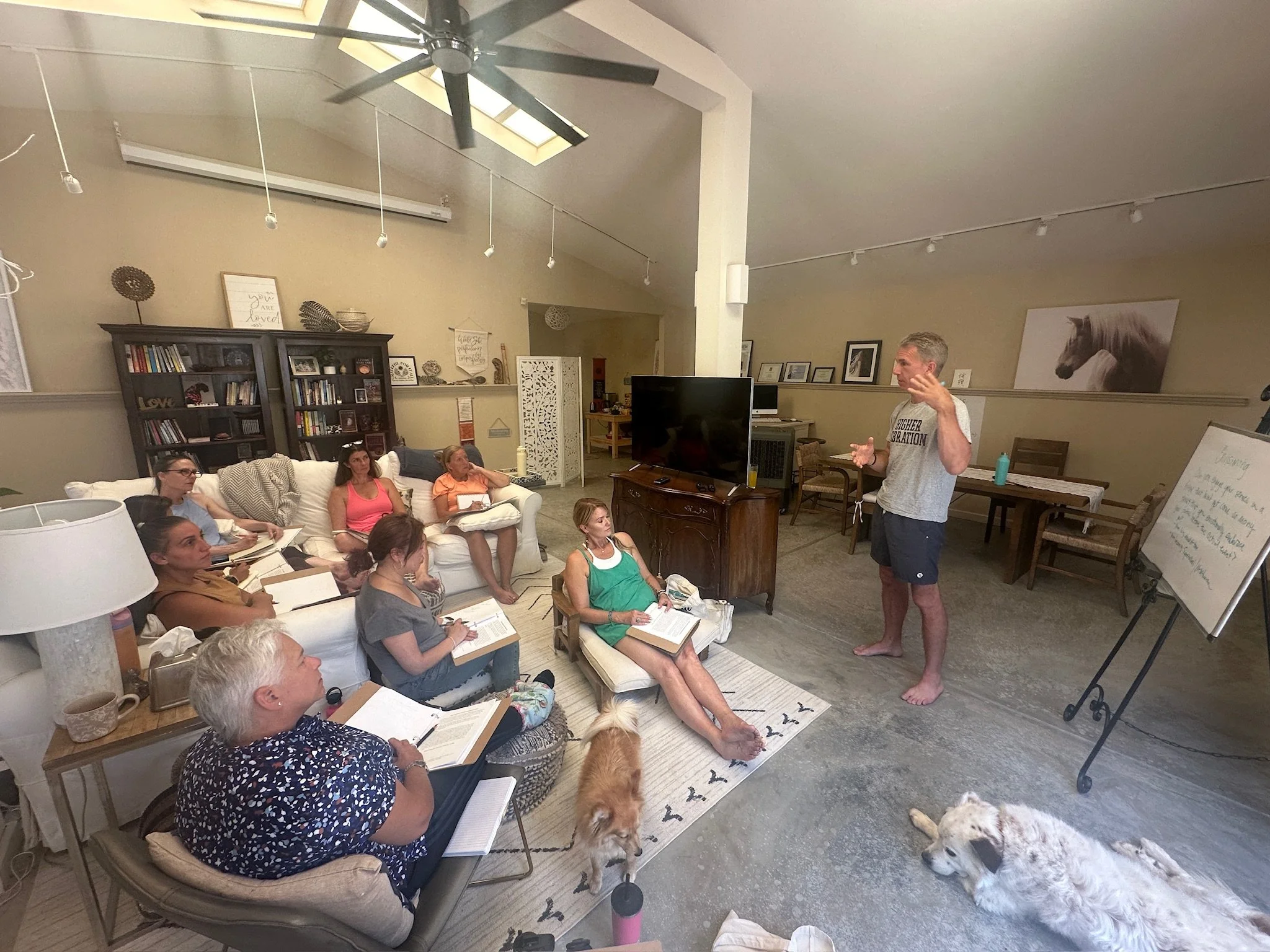 A group of people attending a presentation or workshop in a cozy living room setting with a white sofa, bookshelves, and framed artwork. The speaker, a man in casual clothing, is standing barefoot near a whiteboard, engaging the audience. Two dogs ar