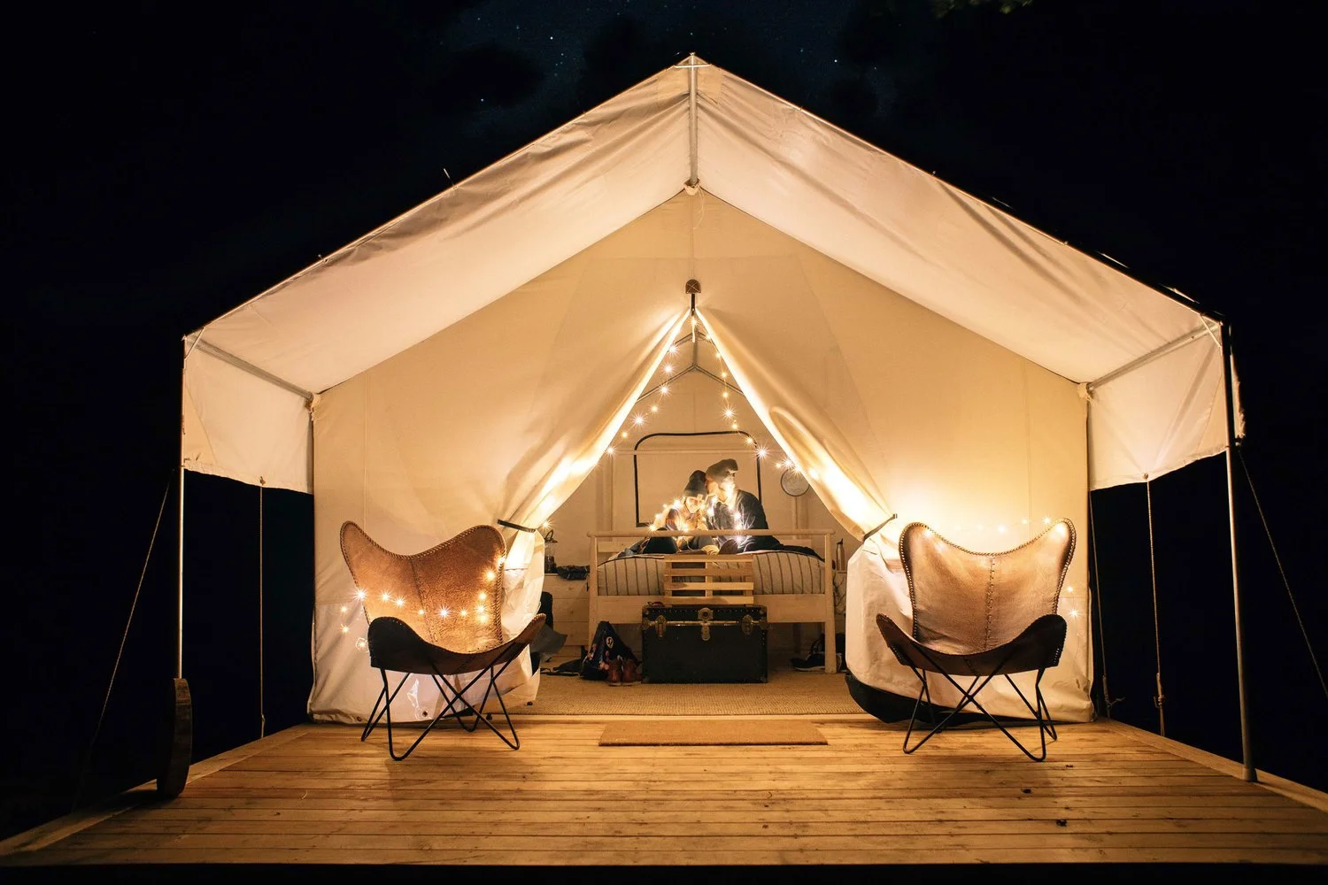 A cozy, illuminated tent set up on a wooden deck at night, with two people sitting inside at a table, surrounded by fairy lights, two lounge chairs outside, and a dark night sky overhead.