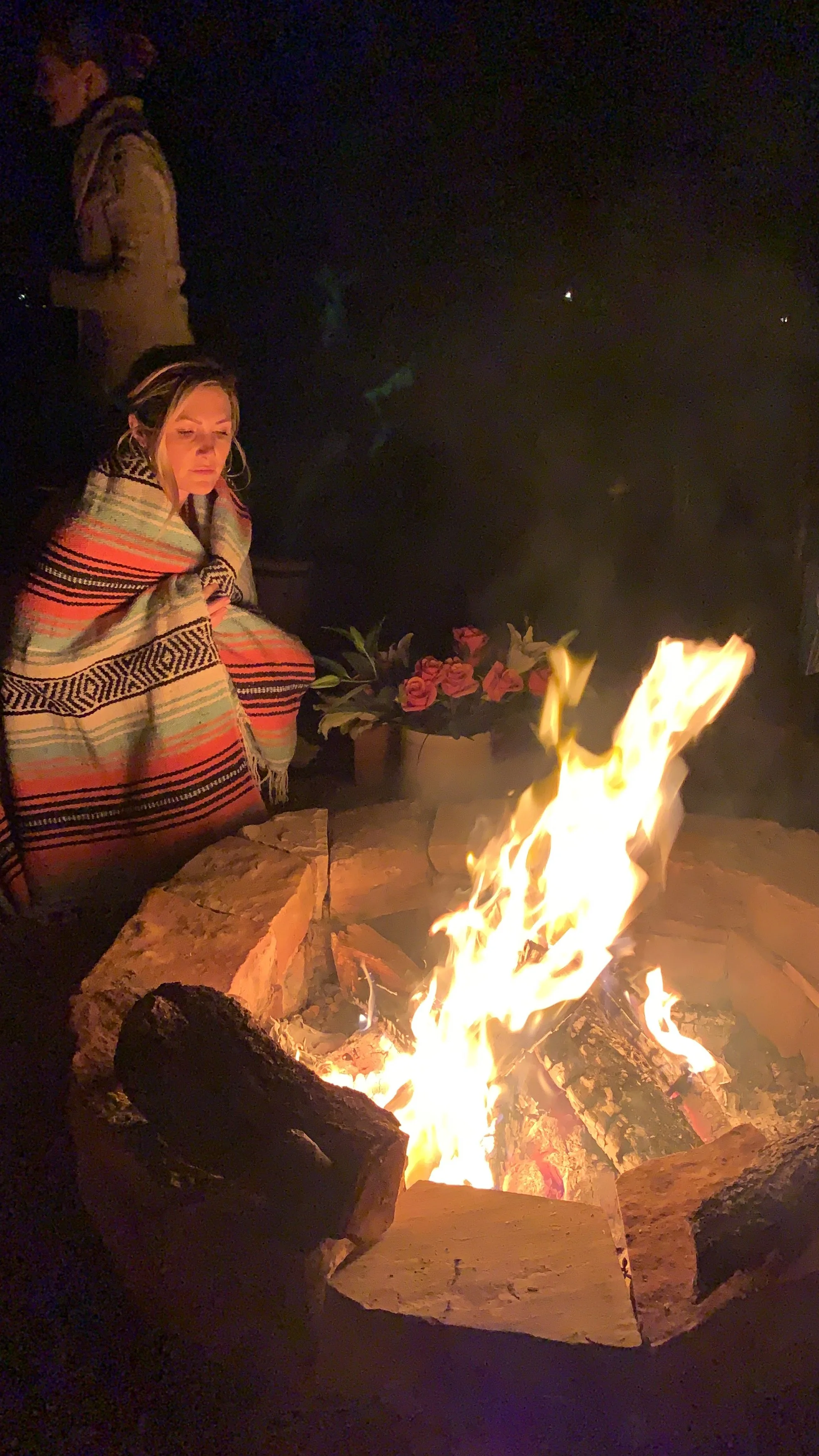 Person wrapped in a colorful blanket sitting by a campfire at night, with another person standing in the background and a pot of flowers nearby.