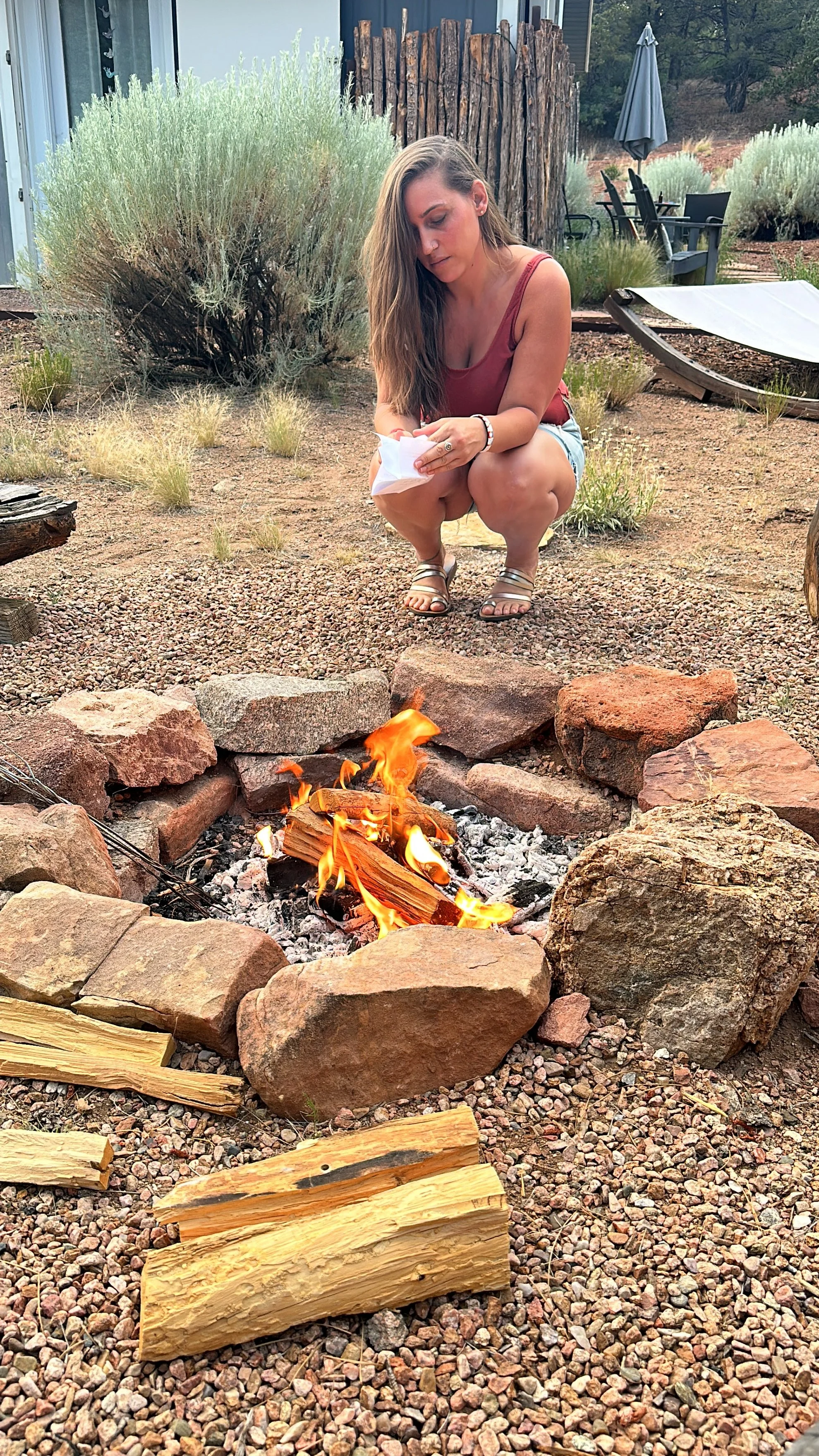 A woman squatting by a fire pit made of large rocks, with small wood pieces burning inside, as she looks at a piece of paper. The background shows outdoor furniture, an umbrella, and landscaped bushes.