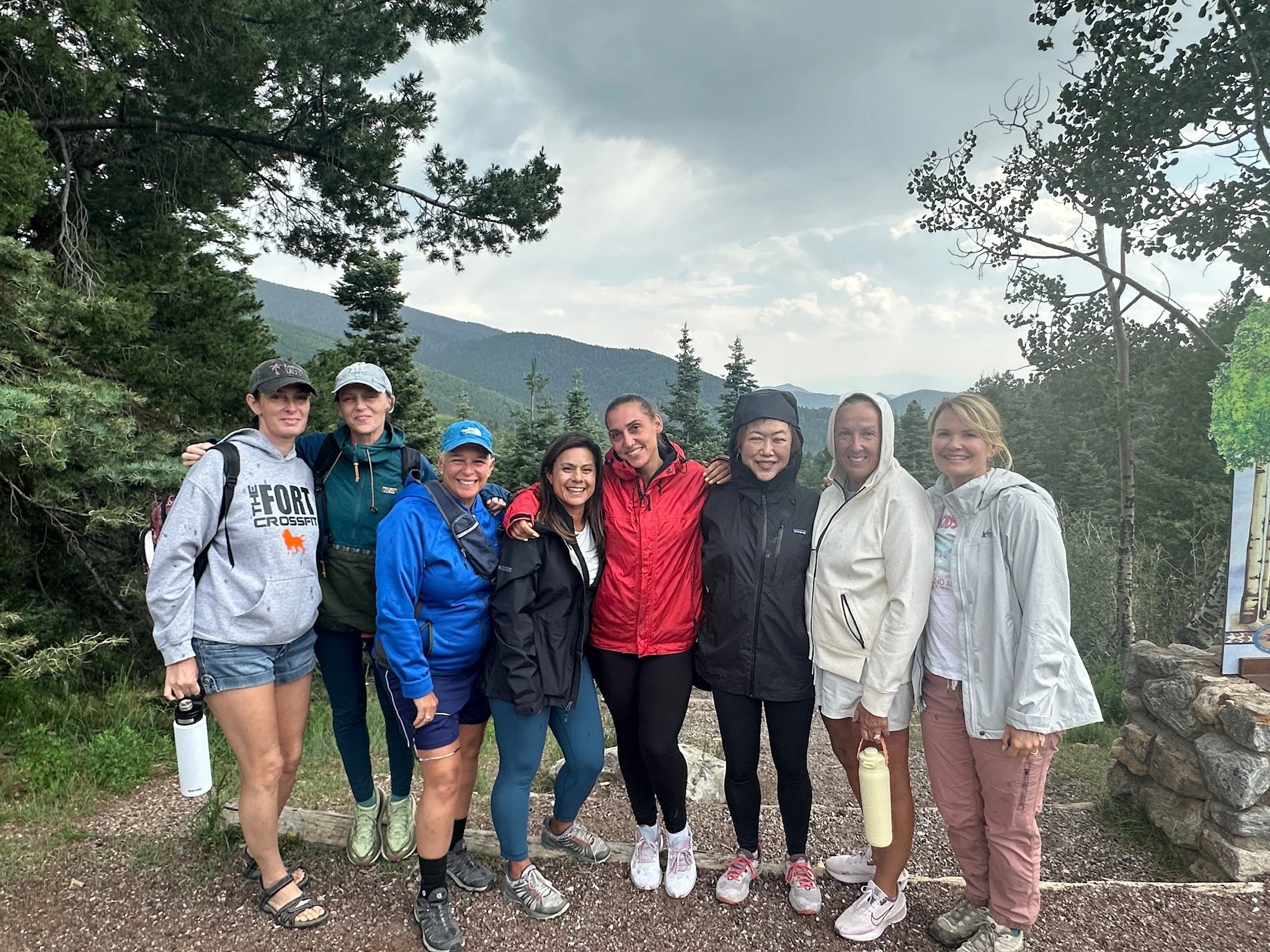 A group of ten women standing outdoors on a trail in a forested mountainous area, smiling at the camera, dressed in outdoor hiking attire, with trees, mountain peaks, and cloudy sky in the background.