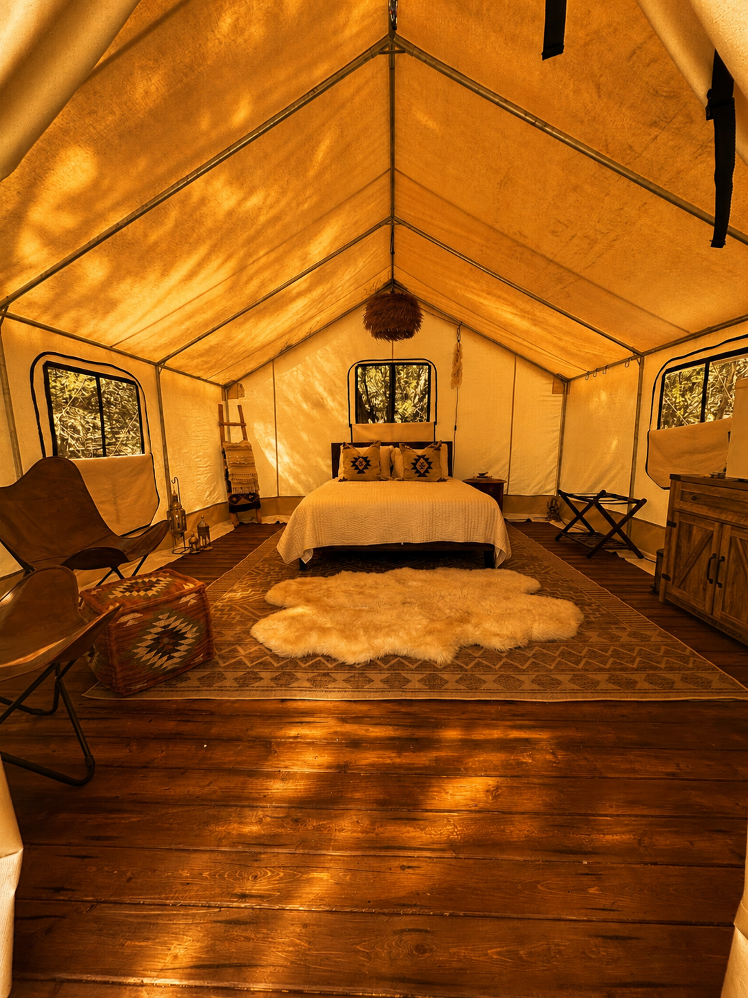 A cozy, illuminated tent set up on a wooden deck at night, with two people sitting inside at a table, surrounded by fairy lights, two lounge chairs outside, and a dark night sky overhead.