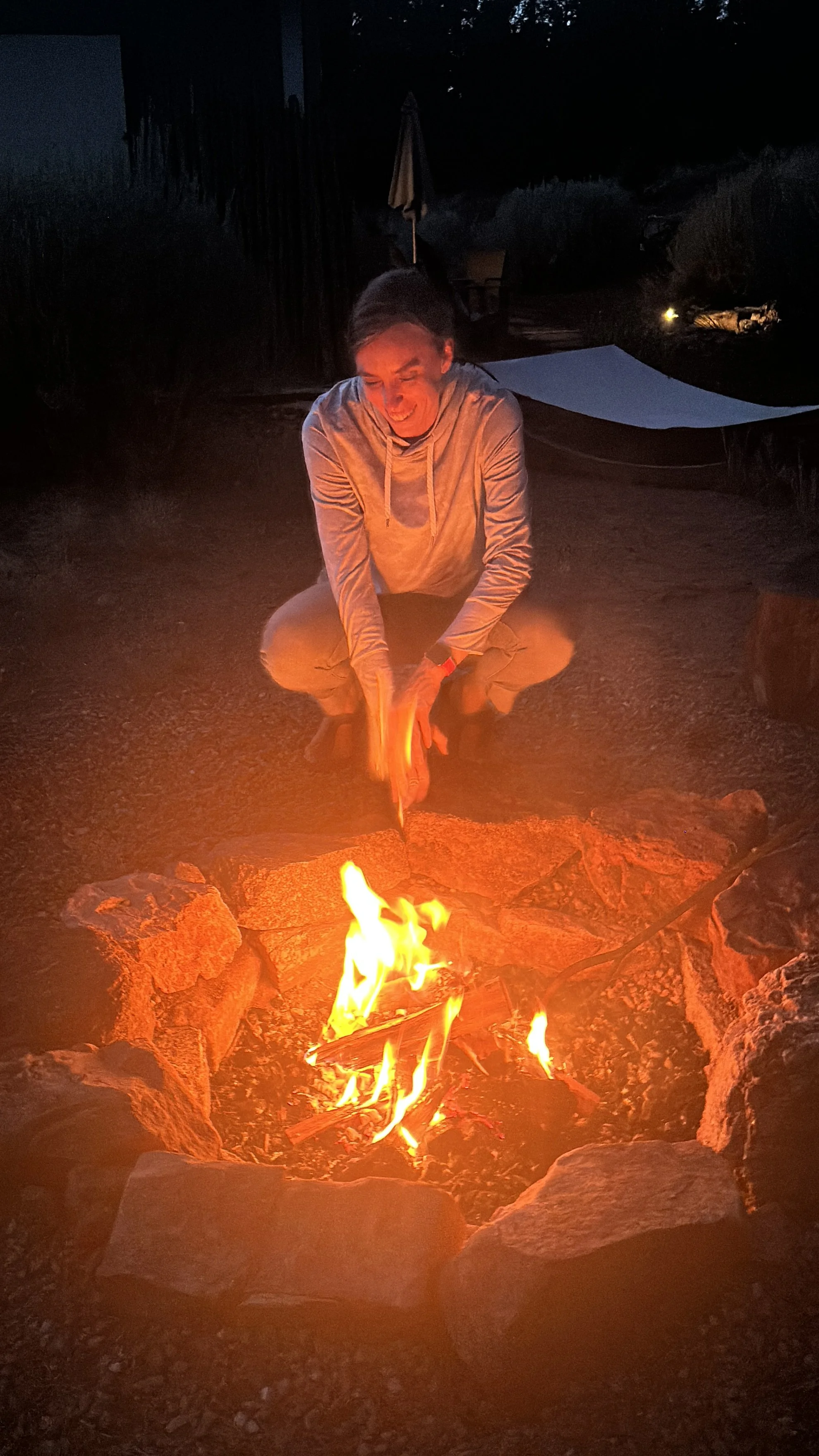 Person crouching by a campfire at night, tending to it outdoors with rocks forming a fire circle.