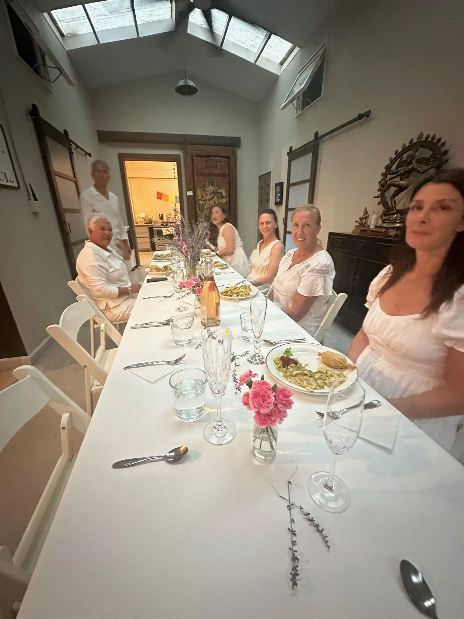 A group of five women seated at a dining table, smiling, with plates of food, glasses, and pink flowers in vases, in a bright room with skylights.