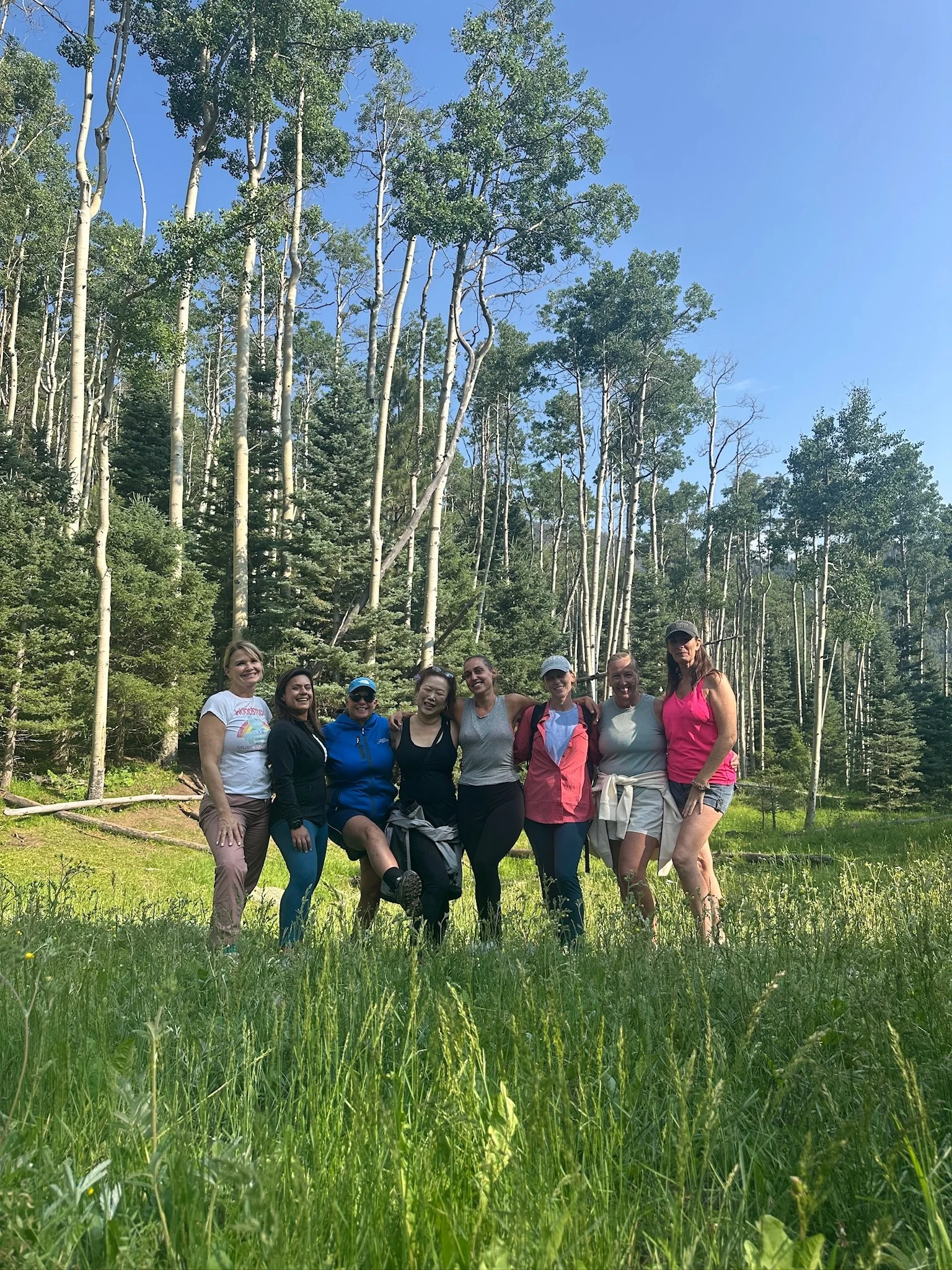A group of eight women standing together outdoors in a grassy area with tall trees and blue sky in the background. They are smiling and posing for the photo, dressed in casual outdoor clothing.