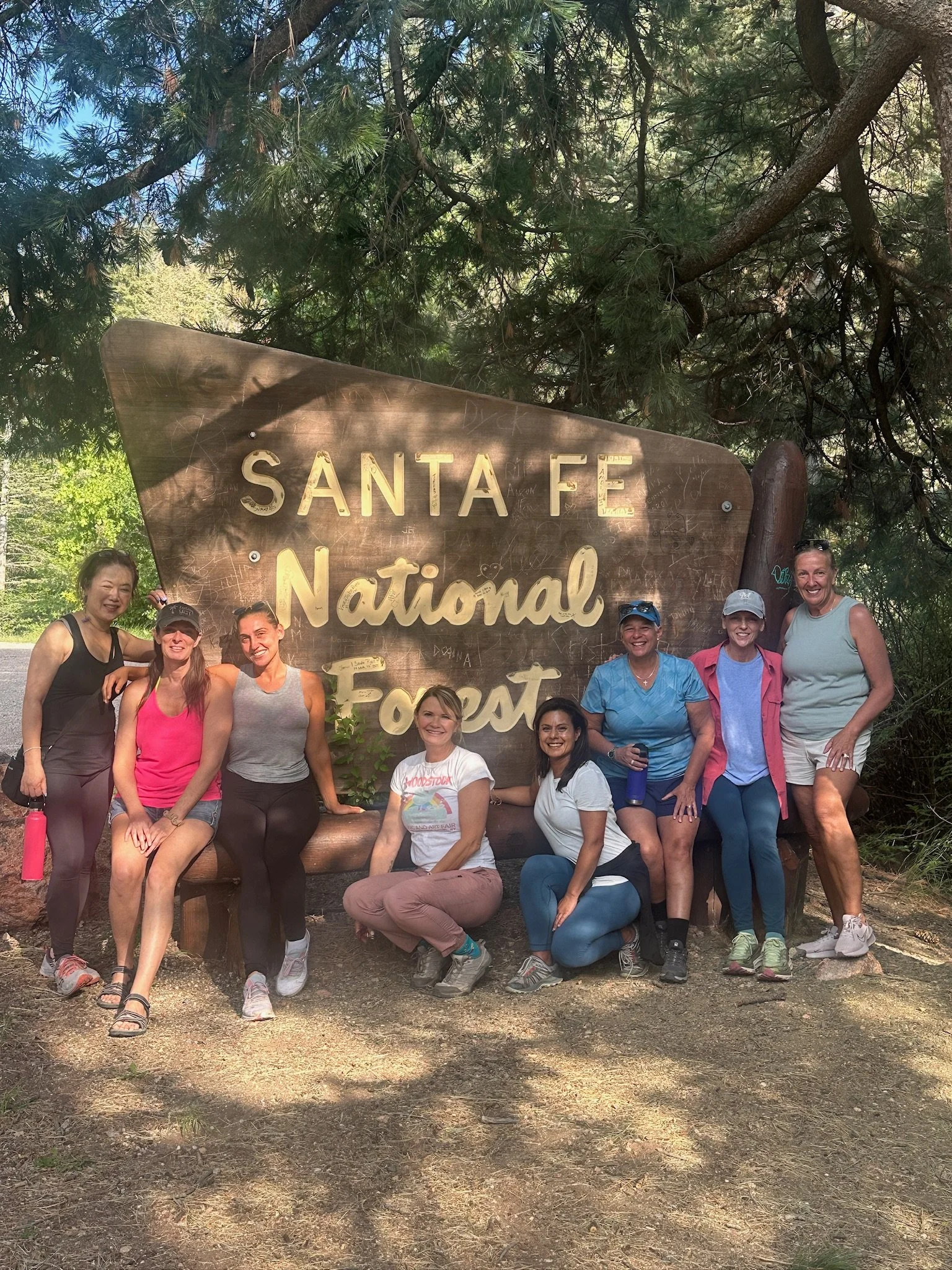 Group of women posing in front of a wooden sign that reads 'Santa Fe National Forest' in a forest setting.