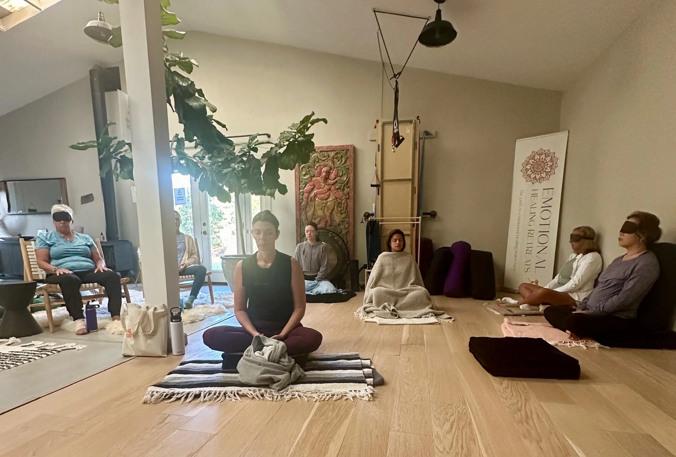 People practicing yoga or meditation in a spacious, well-lit room with plants, artwork, and spiritual decorations.