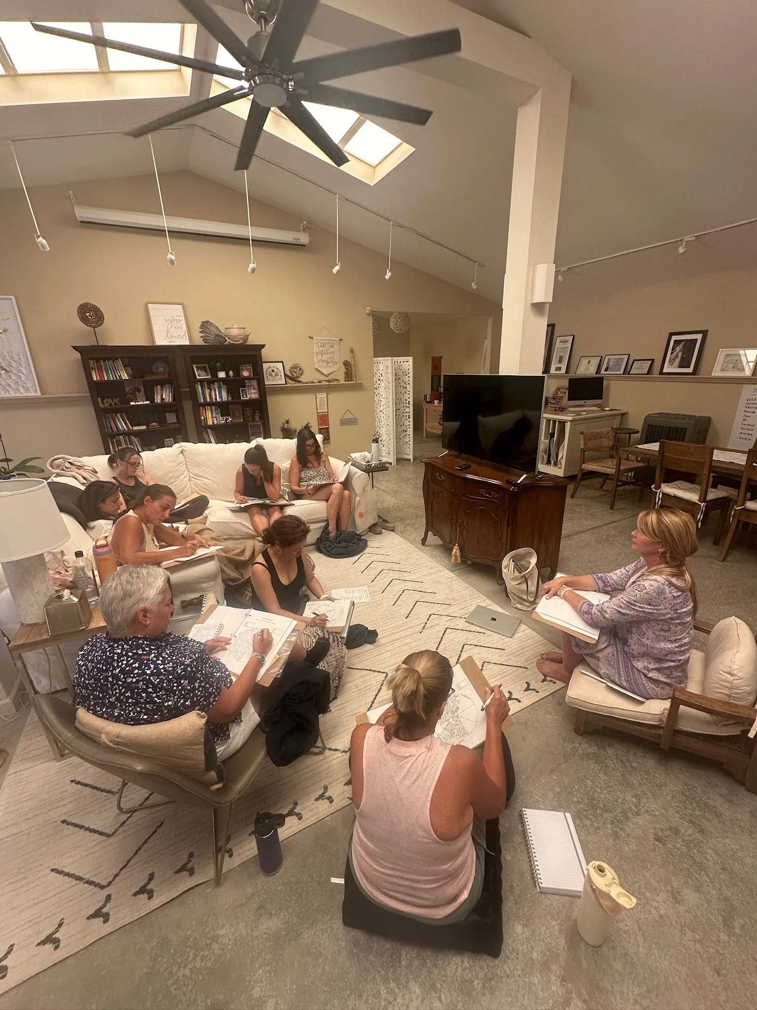 Women sitting on couches and chairs in a living room, engaged in a group discussion or workshop, with notebooks and papers in front of them, and a large TV on a wooden cabinet.