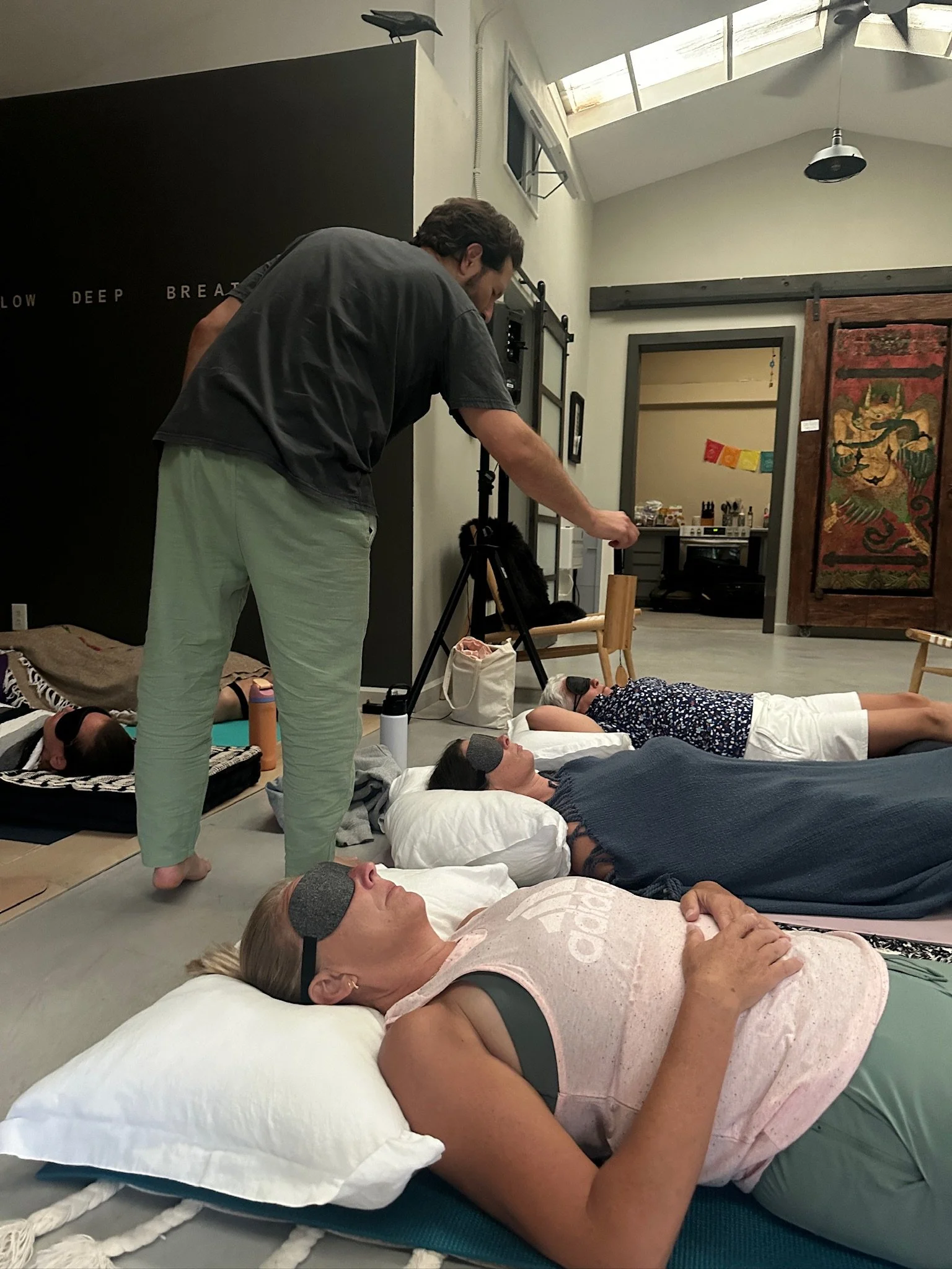 Group of people lying on mats with eyes closed in a yoga or meditation class, some wearing eye masks, in a room with natural light and minimalist decor.