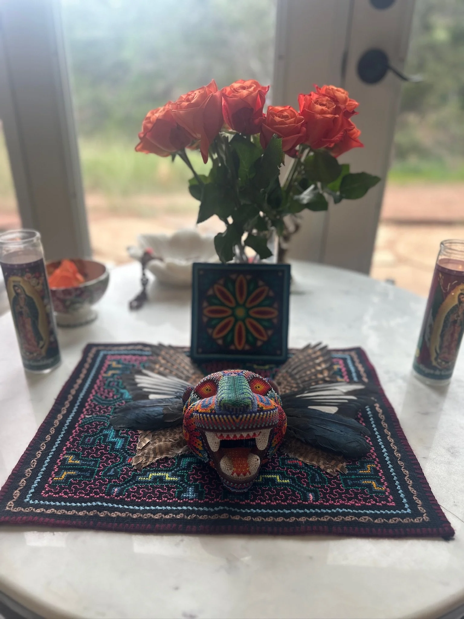 Colorful embroidered skull with wings on a woven base, placed on a decorative cloth on a table, with a blurred background of a window with outdoor greenery.