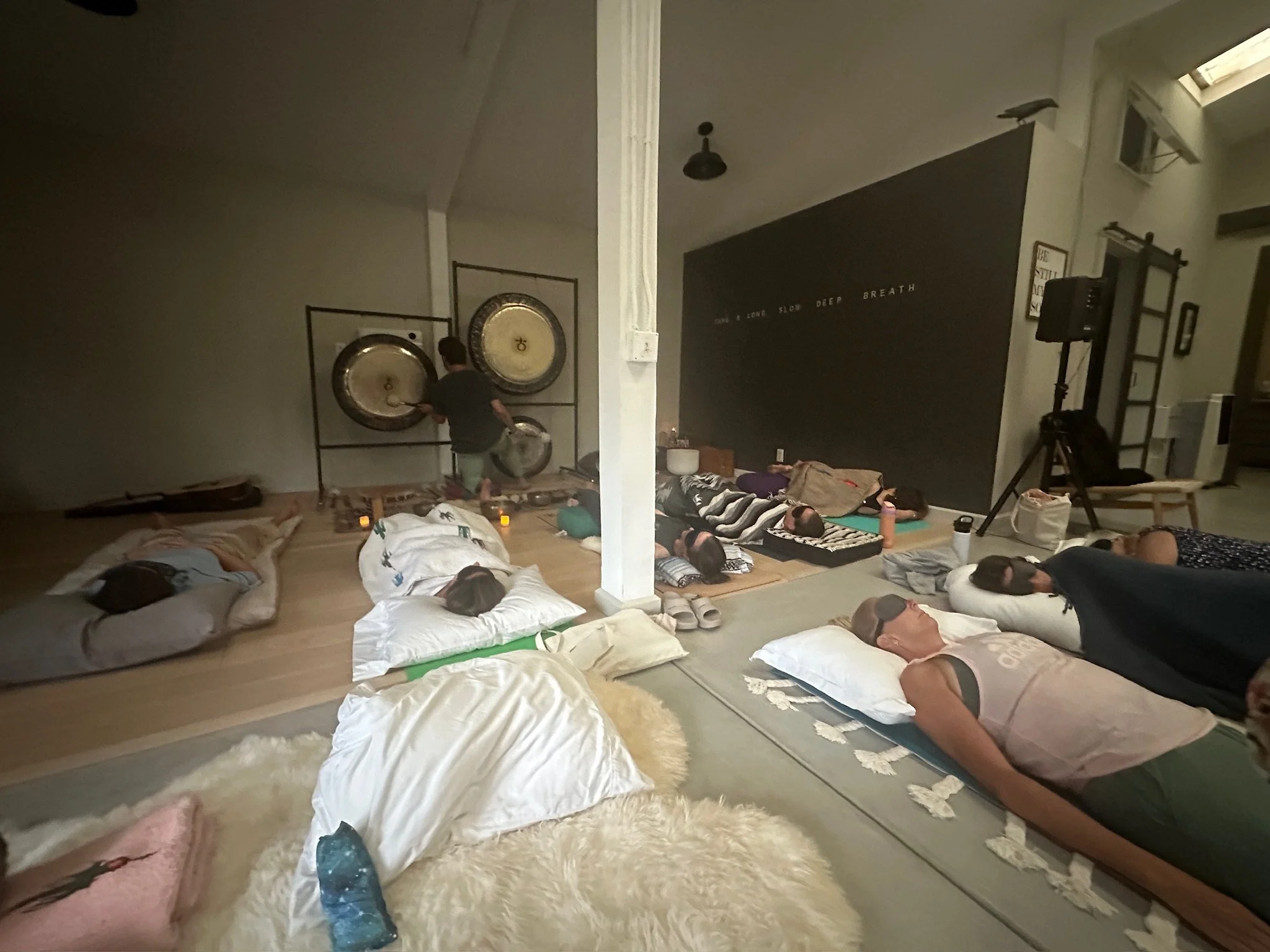 People lying on the floor with pillows and blankets, participating in a group meditation session in a room with a large gong on a stand and a black wall with white writing.