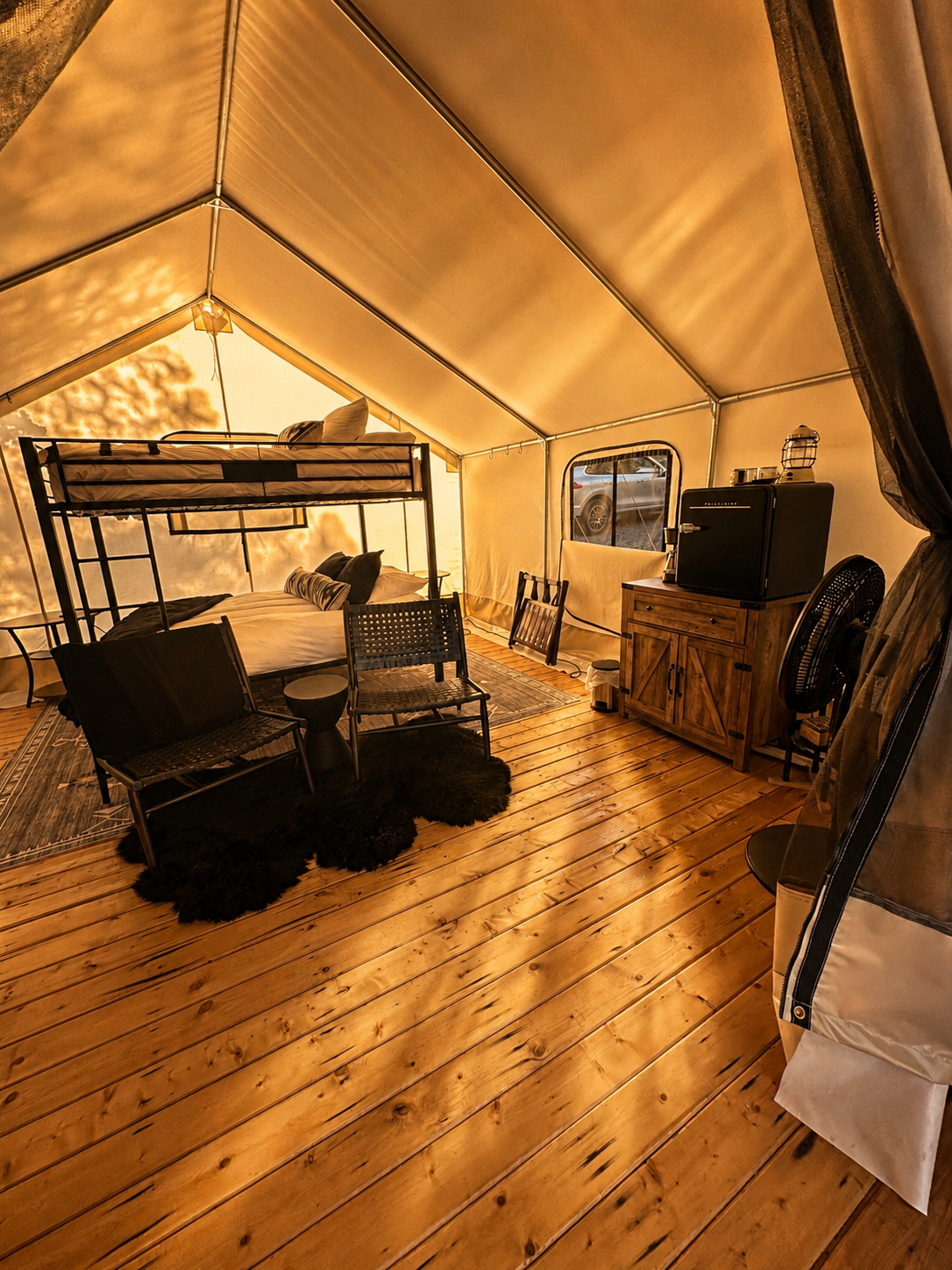 Inside a warm, lit glamping tent with wooden floors, a bunk bed, two chairs with a small table, a cabinet with a fridge, a fan, and a small window showing parked cars outside.