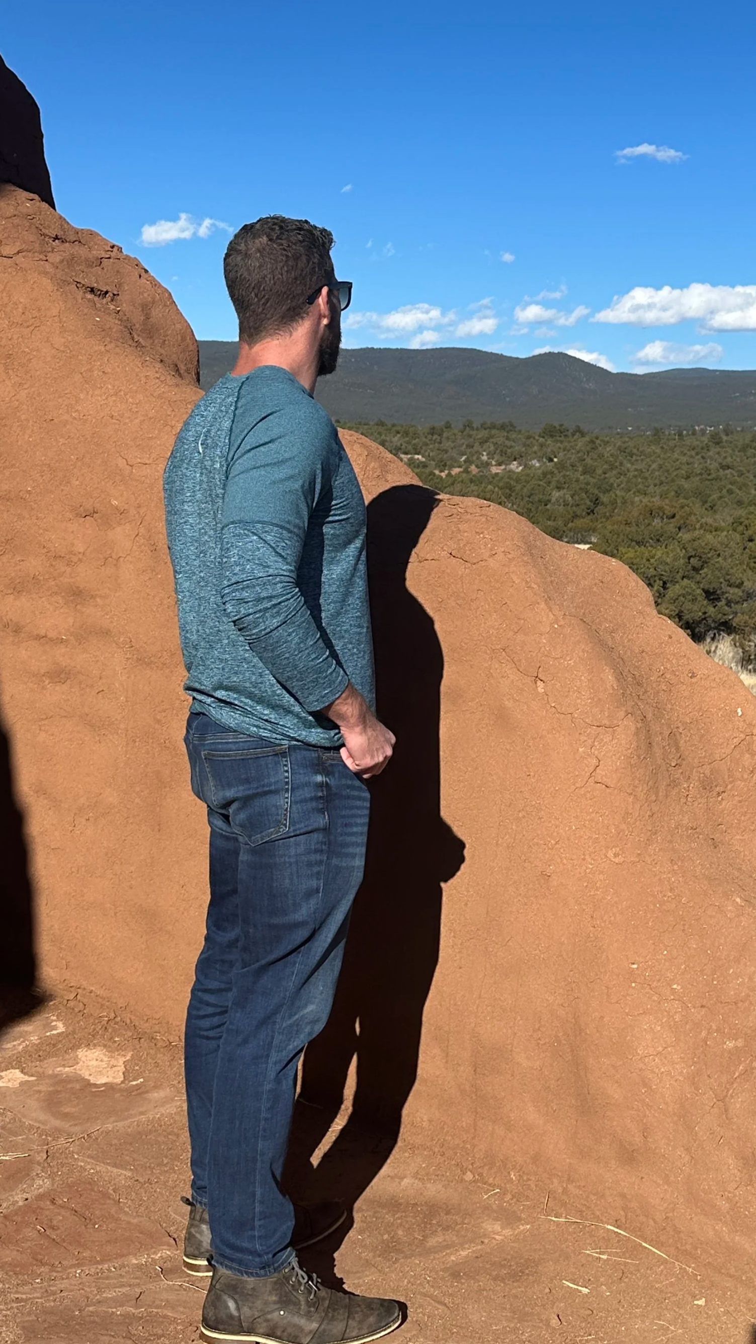 A man with glasses, a beard, and short hair is standing near a large red rock formation, looking into the distance. The background features a clear blue sky with scattered clouds and distant mountain ranges.