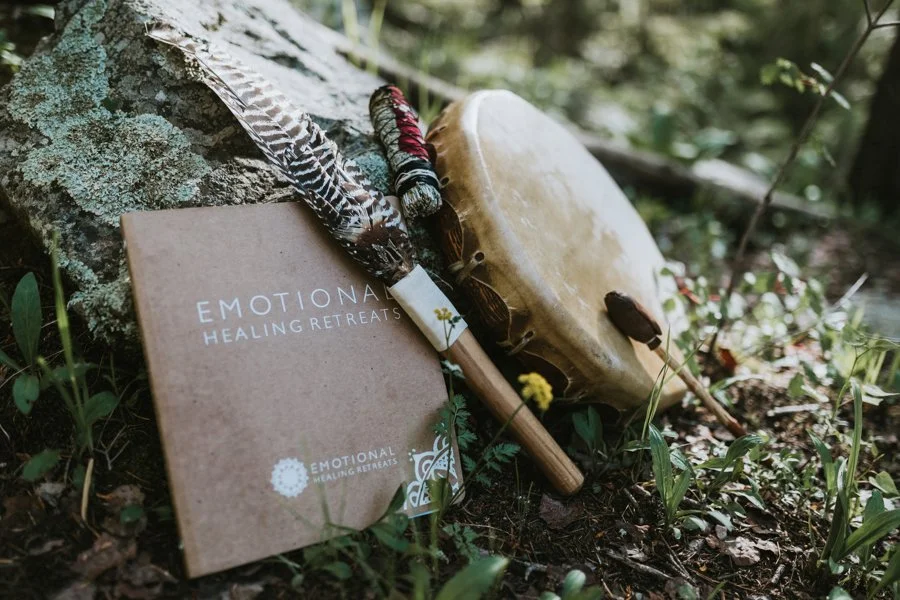 A book titled 'Emotional Healing Retreats' resting on the ground next to a drum, a feather, and a stick with string, surrounded by greenery.