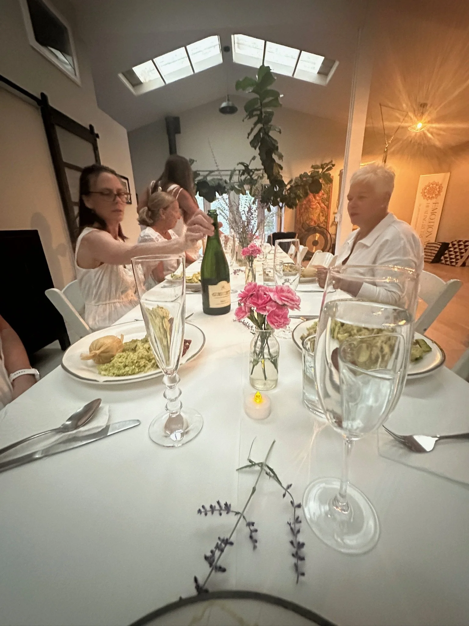 People sitting at a table during a meal in a well-lit dining area with skylights, including flowers, glasses, plates, and a bottle of champagne.