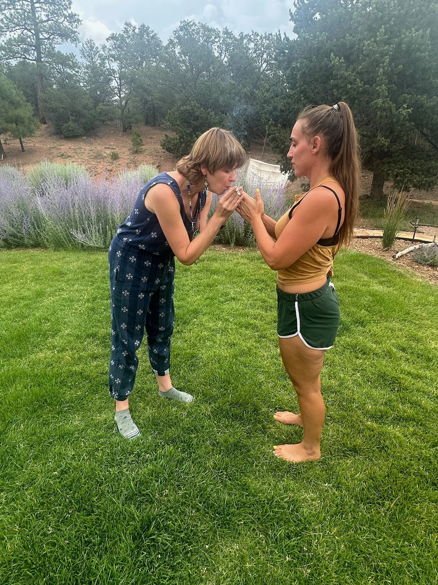 Two women standing on grass outdoors, one woman with short hair wearing a sleeveless blue dress with a patterned design, and the other woman with long hair tied in a high ponytail wearing a yellow and black tank top with green shorts. They are engagi