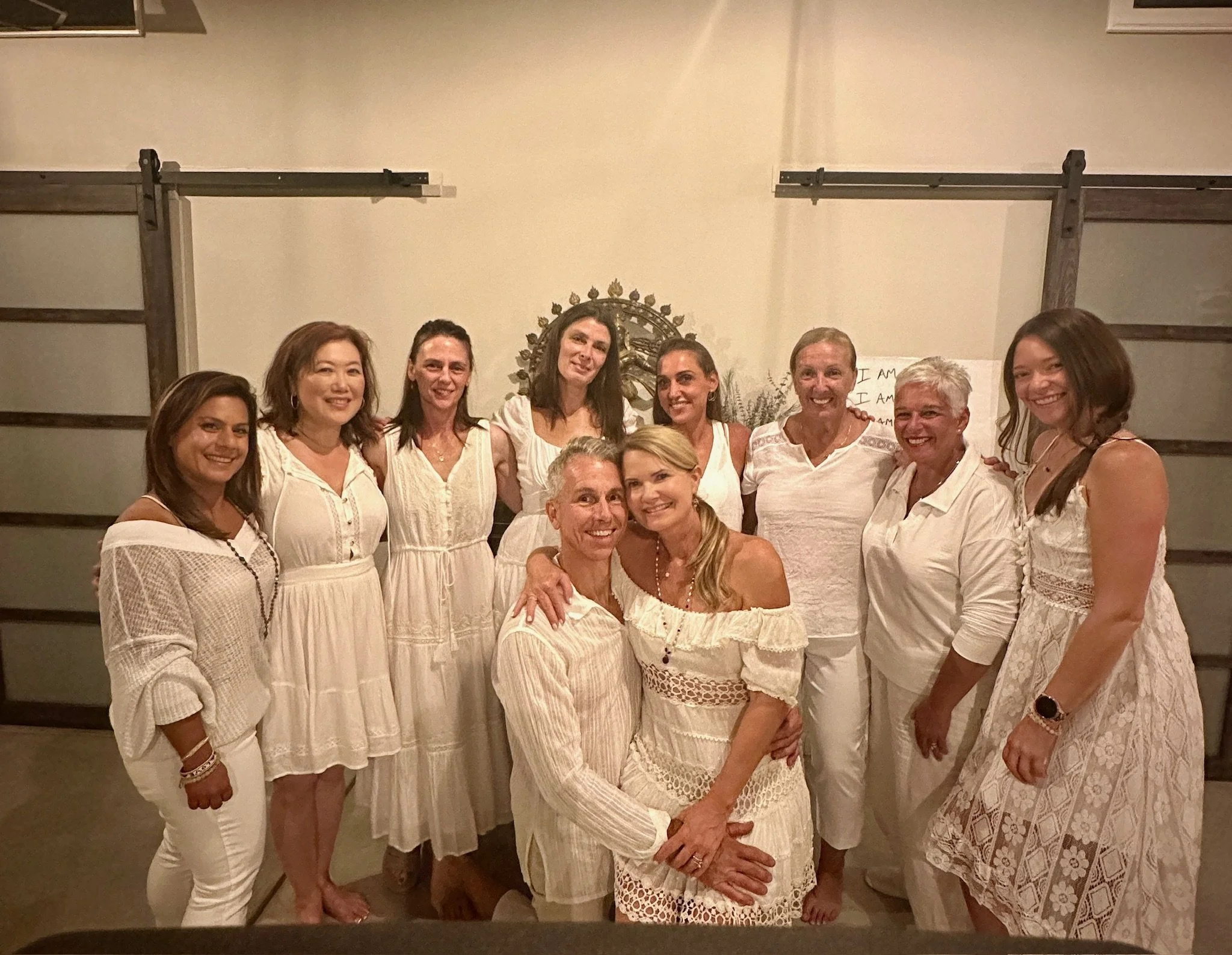 Group of women and one man dressed in white clothing, gathered indoors, smiling and posing for a group photo.