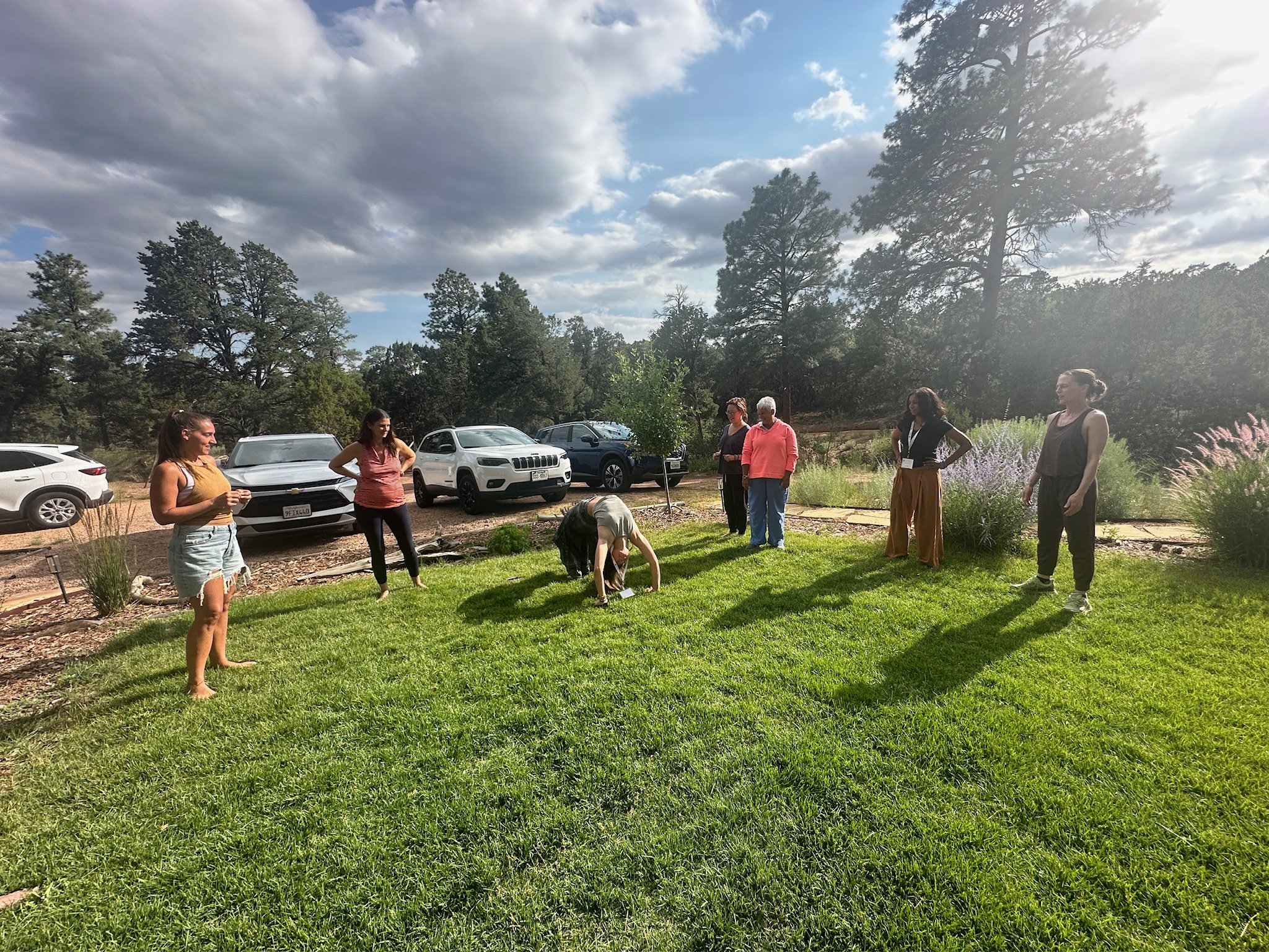 A group of seven women standing outdoors on a grassy area, some chatting and one bending down, with parked cars and tall trees in the background under a partly cloudy sky.