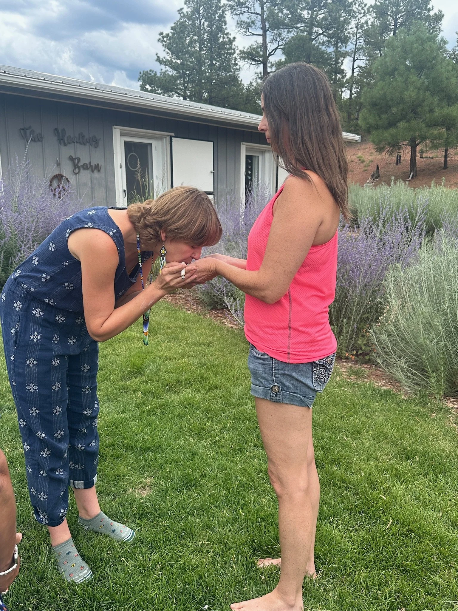 A woman in a pink tank top and denim shorts holds hands with a woman in a blue dress and colorful earrings, who is kissing her hand outdoors in a garden with purple flowers and trees in the background.