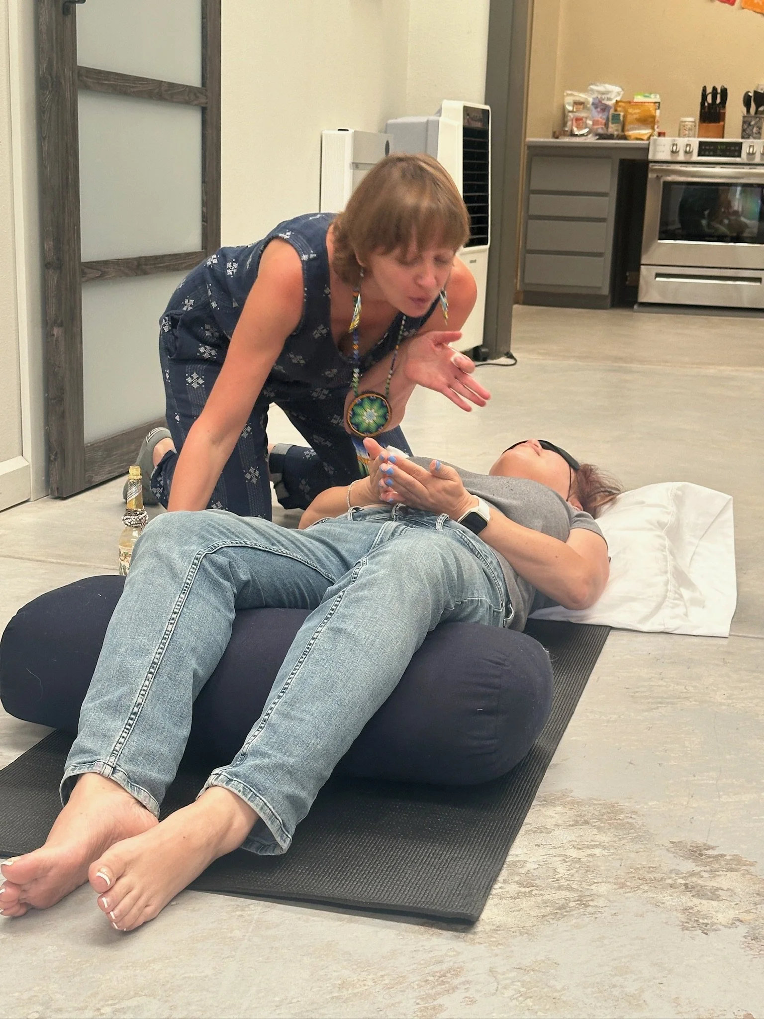A woman wearing glasses, lying on a cushioned mat, appears to be unconscious or sleeping, while another woman kneels beside her, leaning over and possibly tending to her. They are in a home kitchen with appliances and food items in the background.