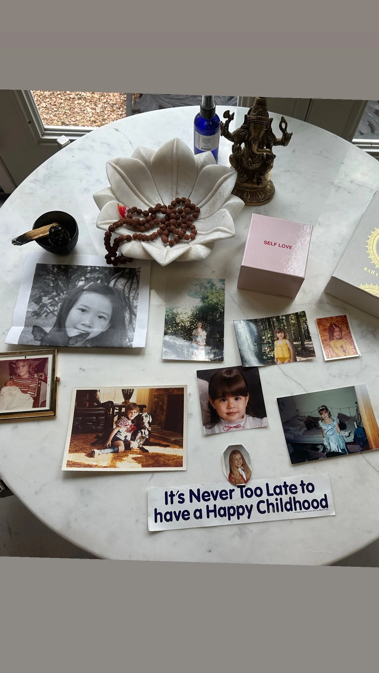 A round marble table displaying a variety of photographs of children, a peace lily flower-shaped dish with a beaded necklace, a small black container with a stick and a black ball, a blue bottle, a brass Ganesha statue, a pink box labeled "Self Love,