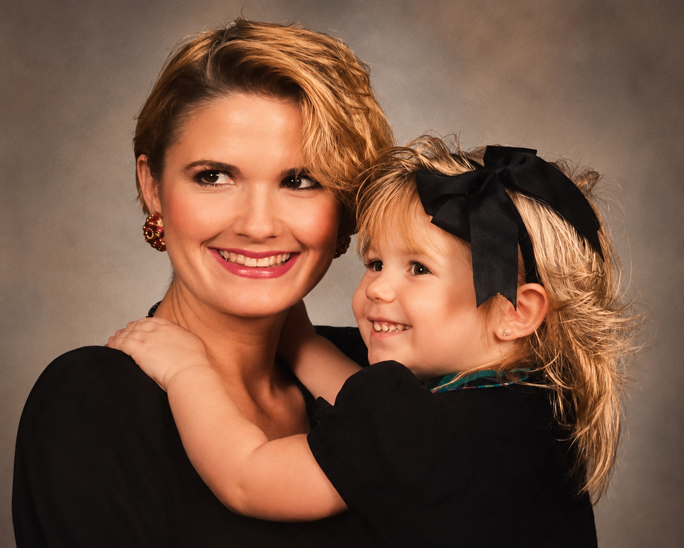 A woman with short blonde hair and a young girl with curly blonde hair, black bow, and earrings smiling and hugging each other against a neutral background.