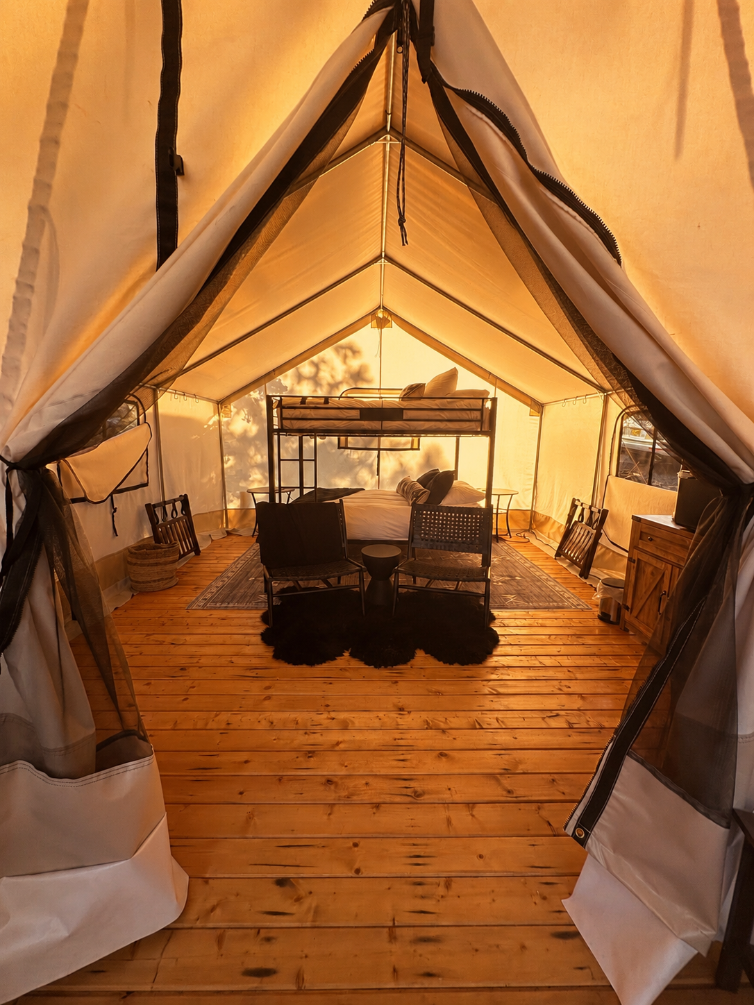 Interior of a tent with a bunk bed, two black chairs, a small black side table with a candle, and a black furry rug on wooden flooring.