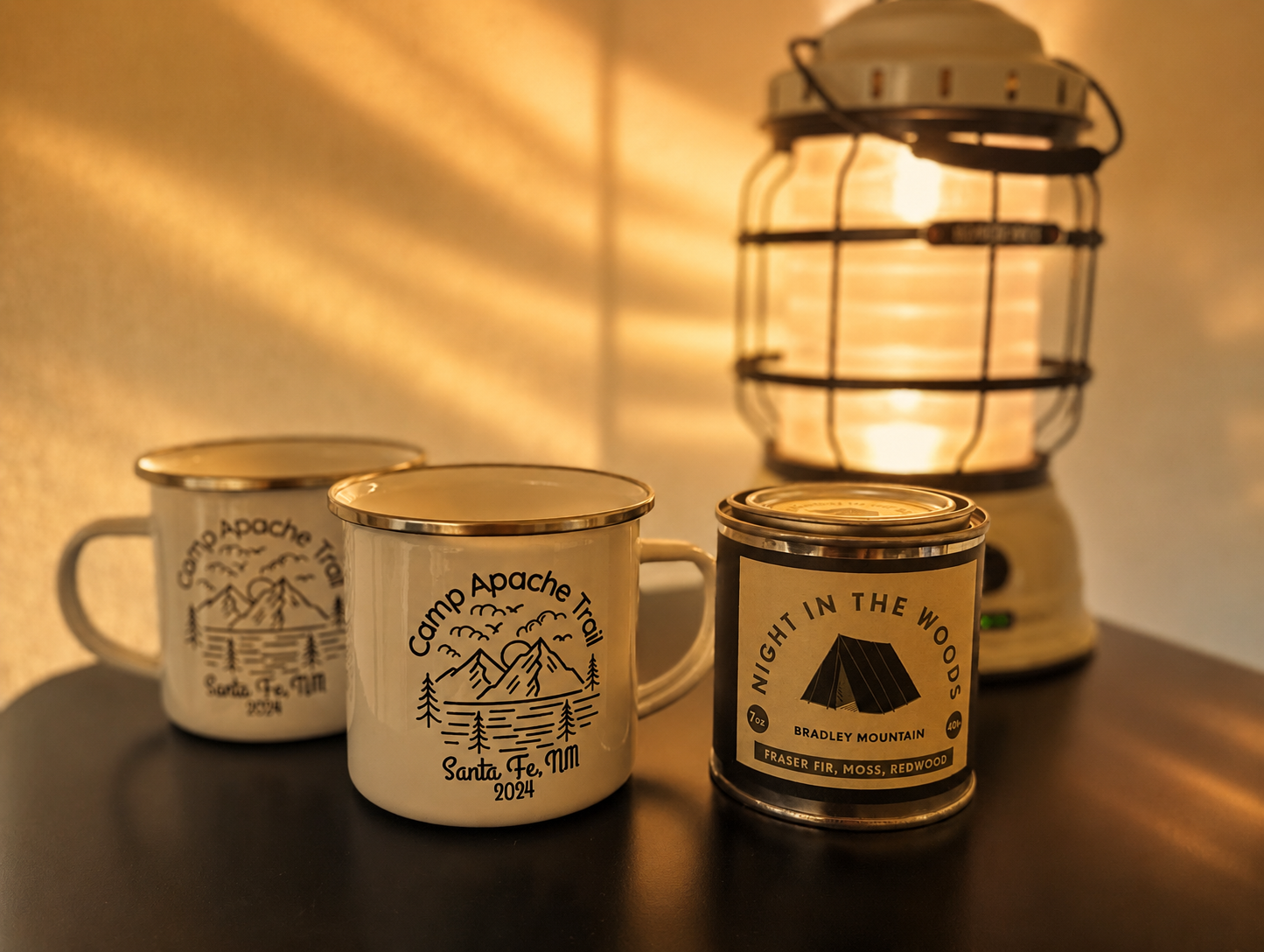Three camping mugs and a candle on a dark surface beside a lantern illuminating the scene with warm light.