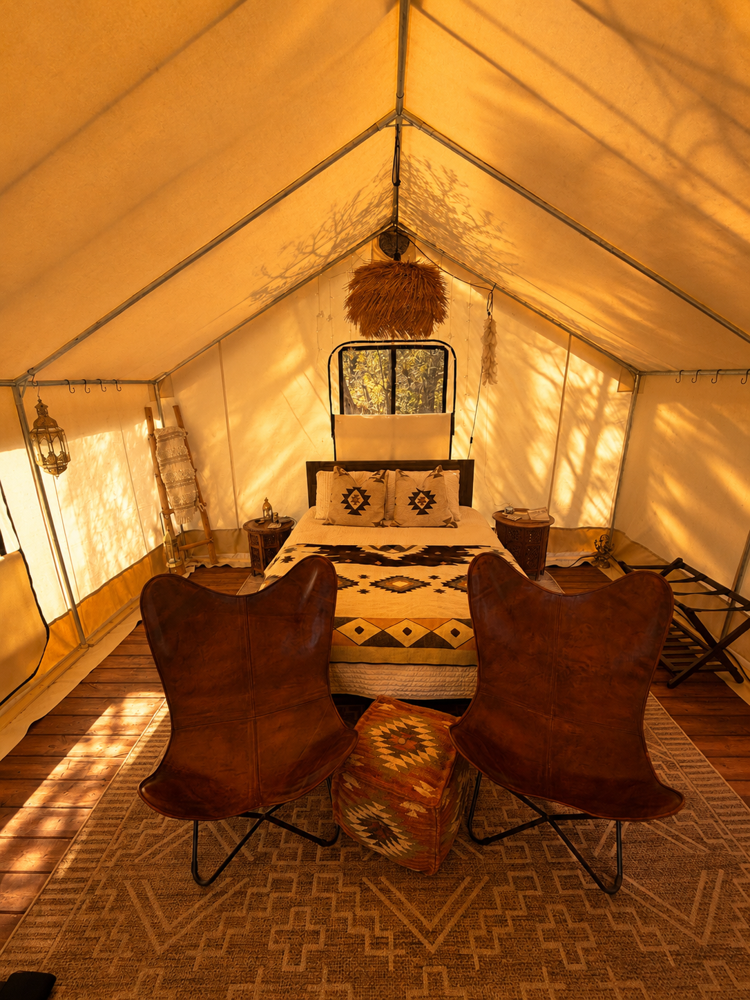 Interior of a cozy, rustic tent with a bed, two armchairs, a rug, and vintage decor, illuminated by warm sunlight.