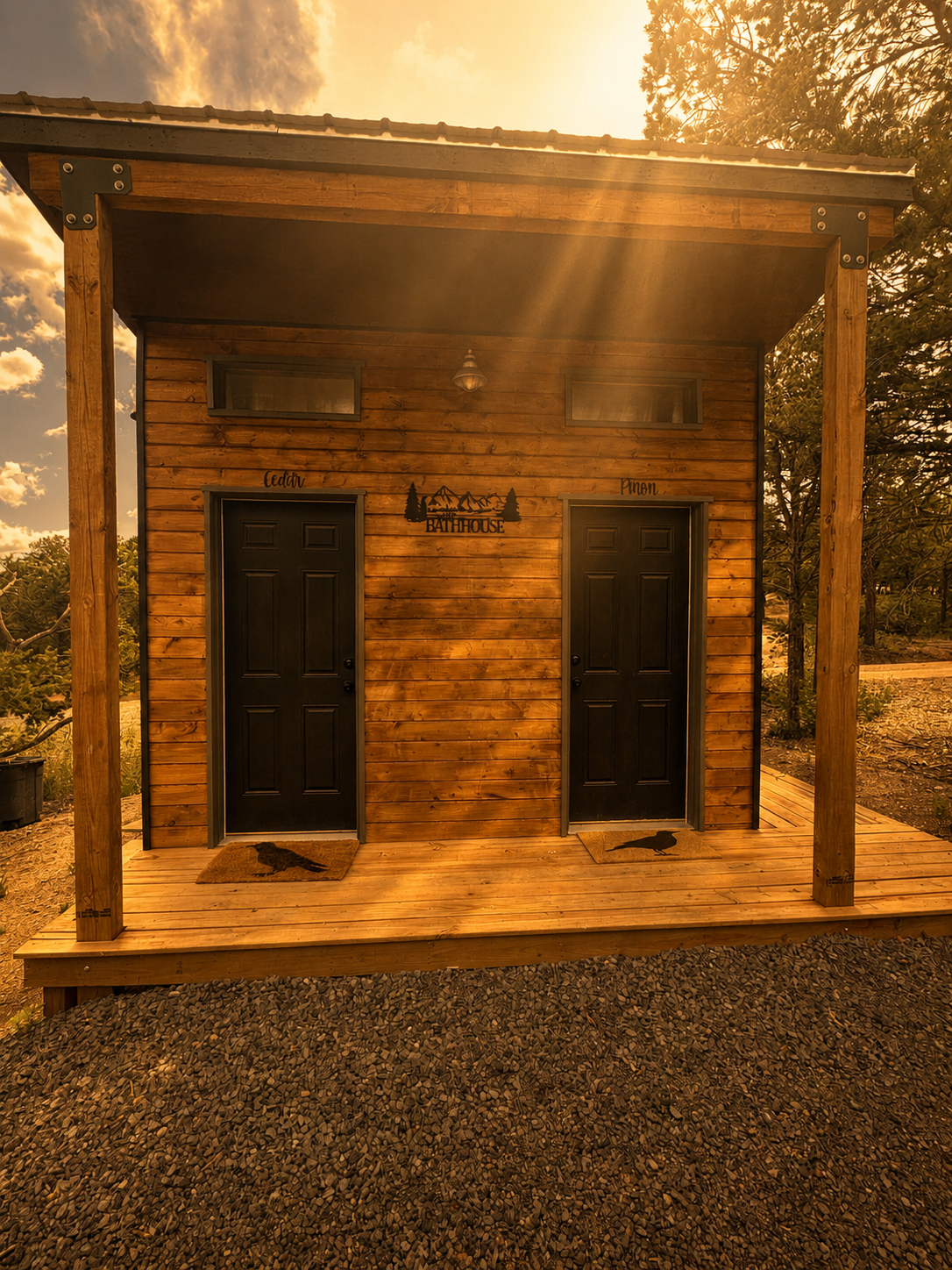 A small wooden building with two black doors, two small horizontal windows, and a porch with two doormats, each with a bird silhouette. The building has a sign that reads "Bathhouse" and is illuminated by warm, golden sunlight.