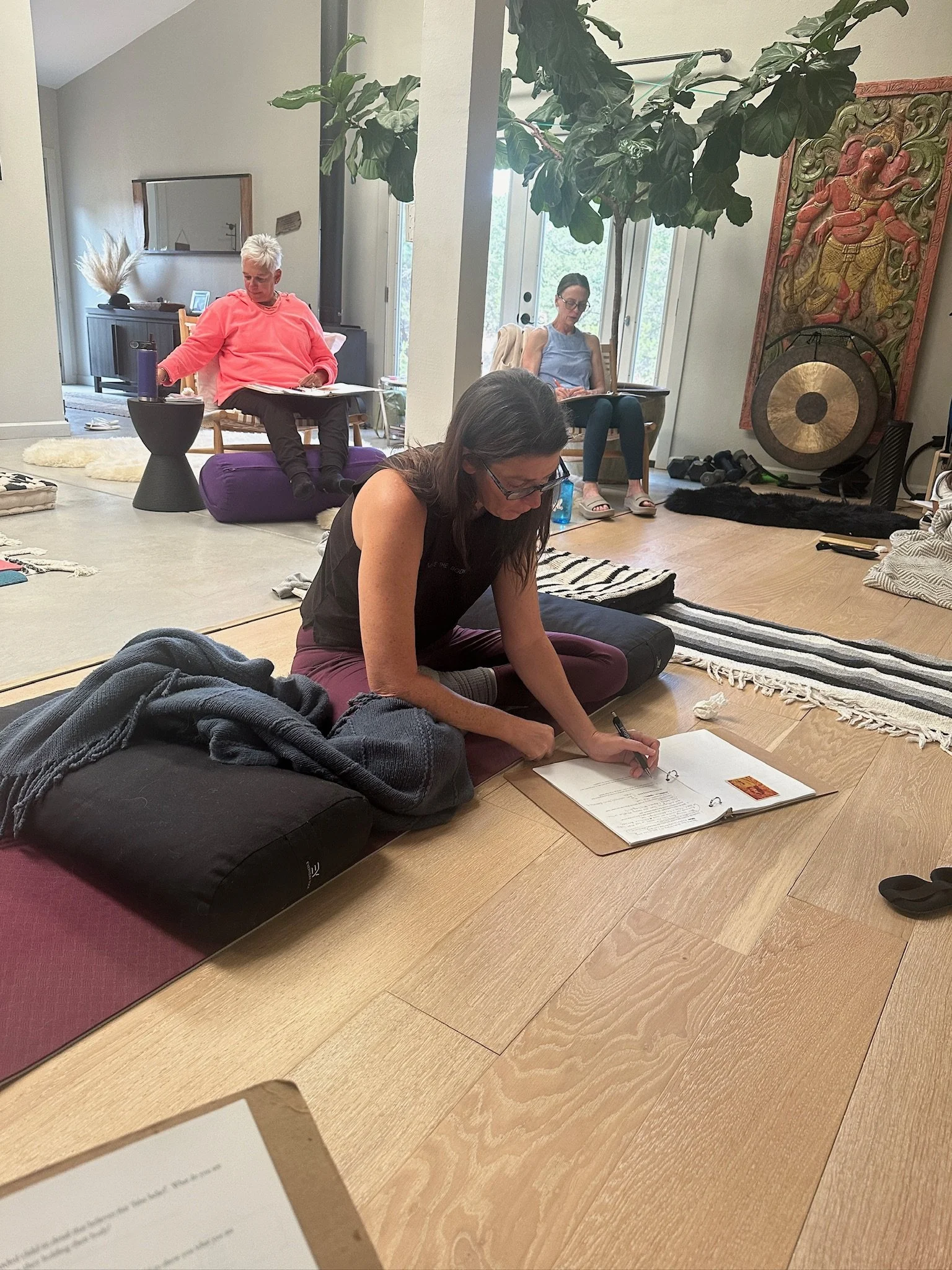 Three women are participating in a yoga or meditation session on the floor of a cozy living room. The woman in the foreground is sitting cross-legged, writing in a notebook, with a blanket and some clothing items nearby. The woman in the background o