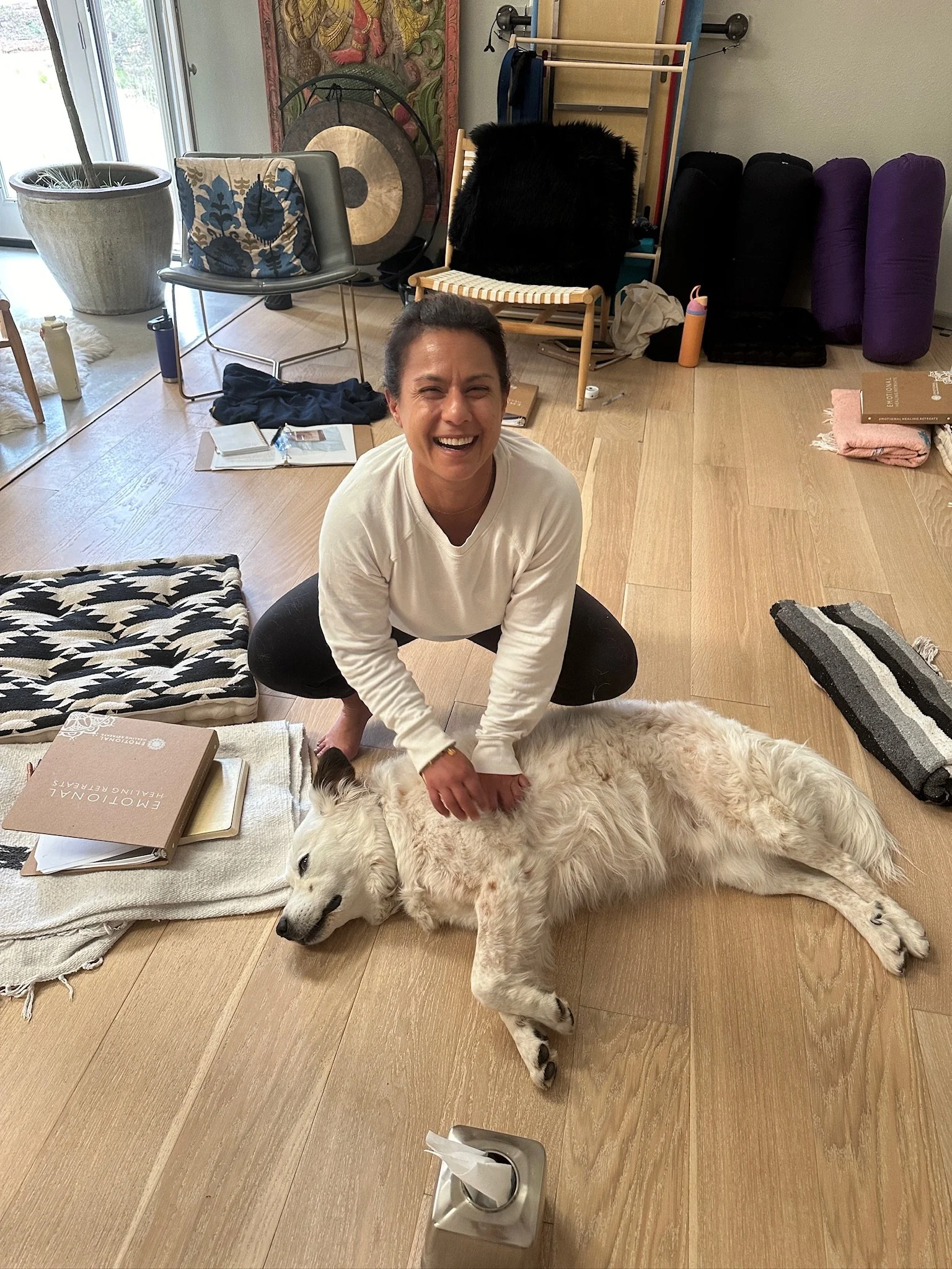 A person squatting and smiling while petting a large, white dog lying on the wooden floor. The person is wearing a white long-sleeve shirt, and the dog appears to be relaxed. The room has various items including a blanket, books, a chair, and yoga ma