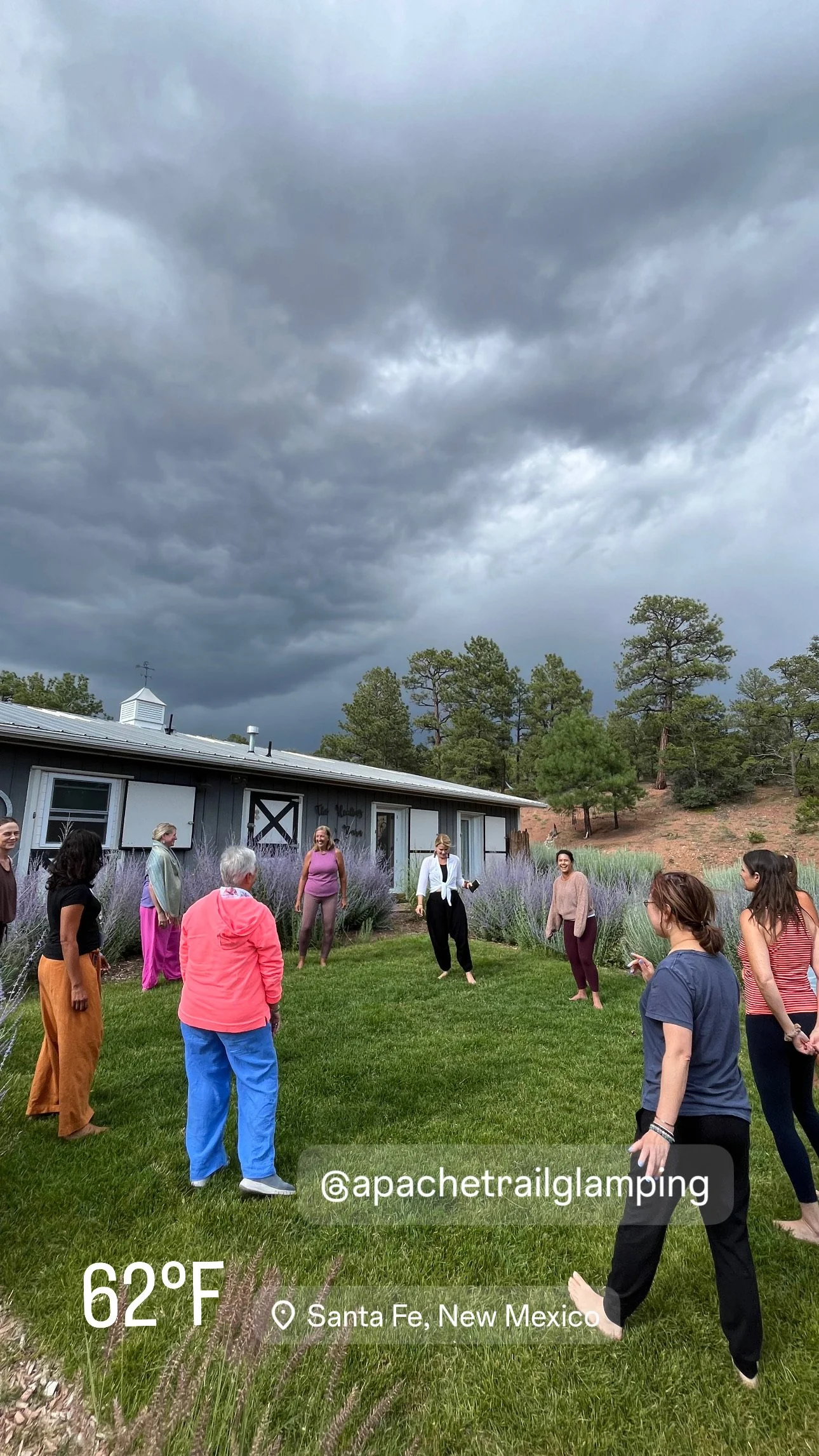 Group of people standing in a circle outside near a gray building and purple flowering plants, with pine trees and dark storm clouds overhead in Santa Fe, New Mexico.