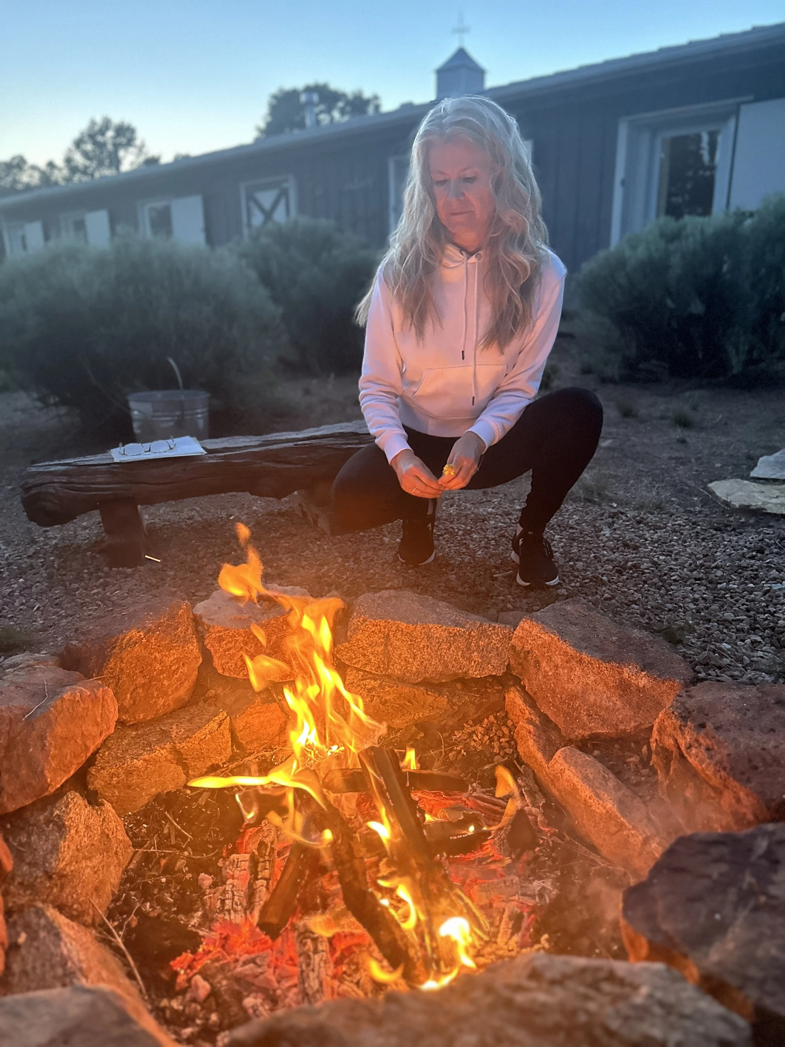 A woman with long blonde hair, wearing a light pink hoodie, sits next to a campfire surrounded by rocks in a rural outdoor setting during evening. Behind her, there is a building with a cross on top and trees in the background.