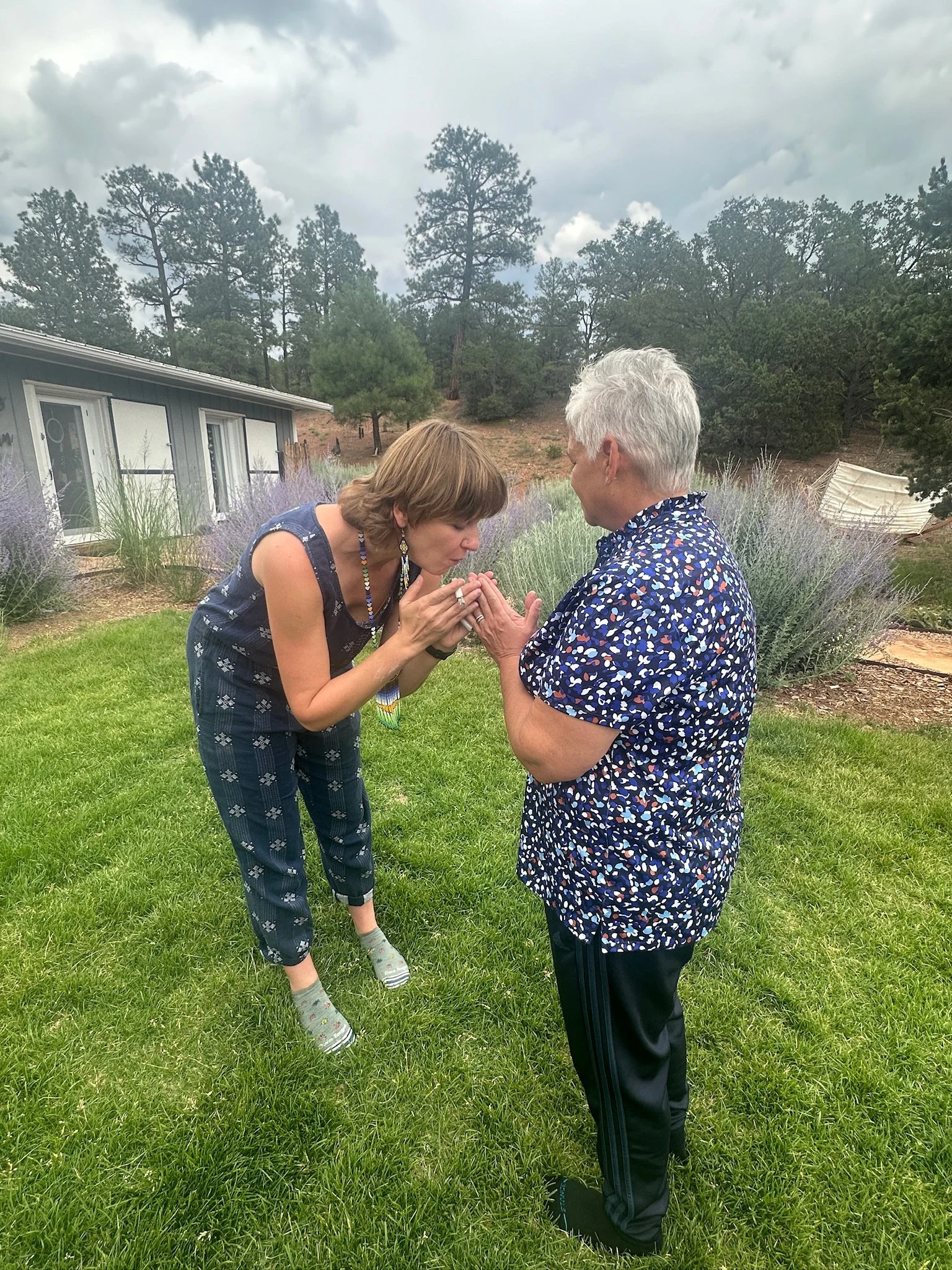 Two women holding hands and leaning towards each other in a grassy outdoor area surrounded by trees, with a house in the background and cloudy sky above.