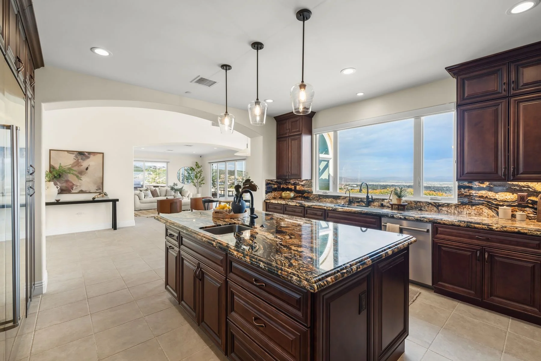 Modern kitchen with dark wood cabinets, a granite countertop, and large windows showing a city view. Recessed lights and three pendant lights hang above the island. In the background, a living room with a white sofa, circular mirror, and potted plant