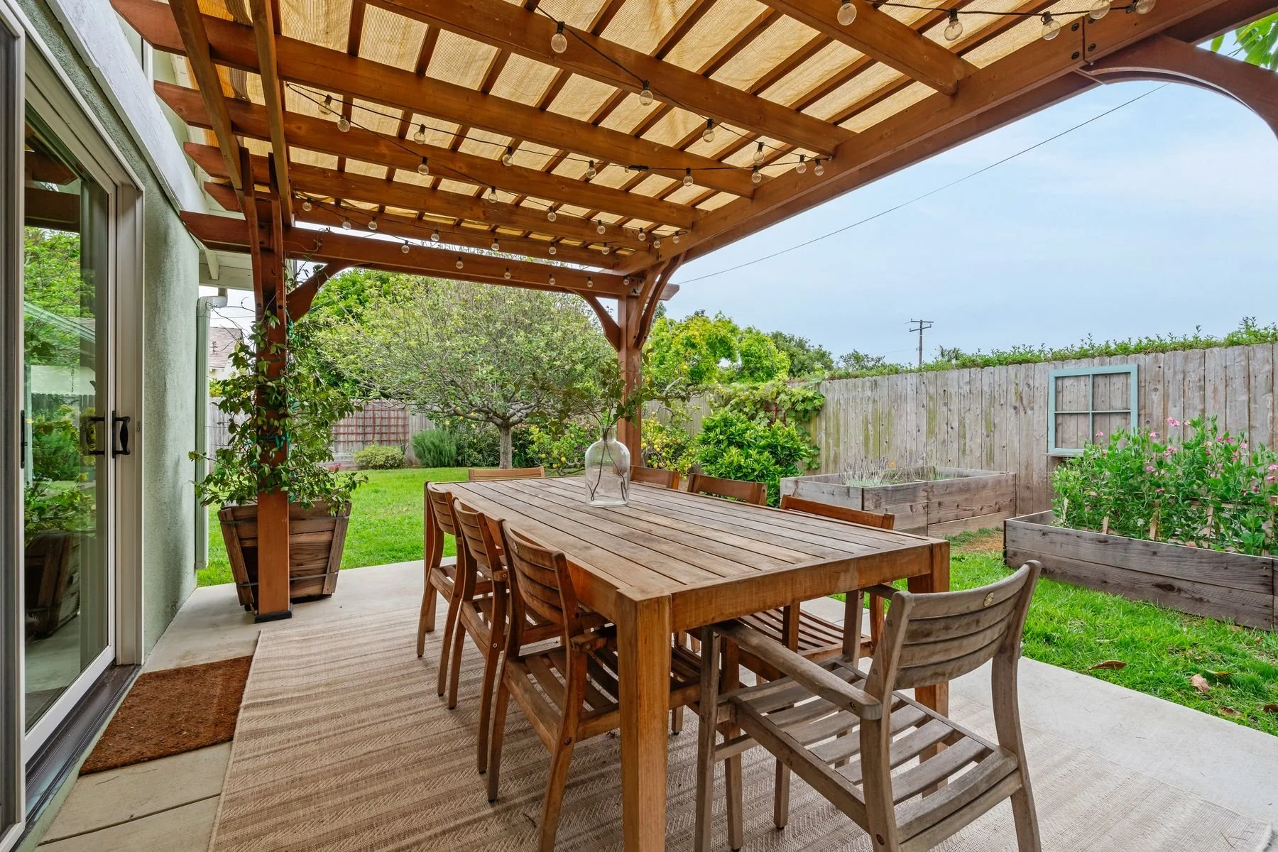 Outdoor patio with wooden table and chairs under a pergola with string lights, surrounded by a green backyard with trees and flower beds.