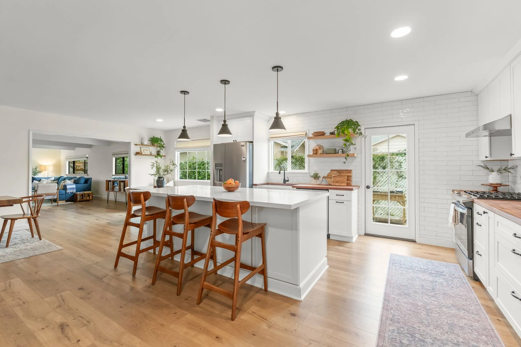 Bright open kitchen with white cabinetry and a large island with seating. Wooden bar stools in front of the island. Stainless steel refrigerator and gas stove. Shelves with plants and decor. Door to outside patio. Adjacent living room with blue couch