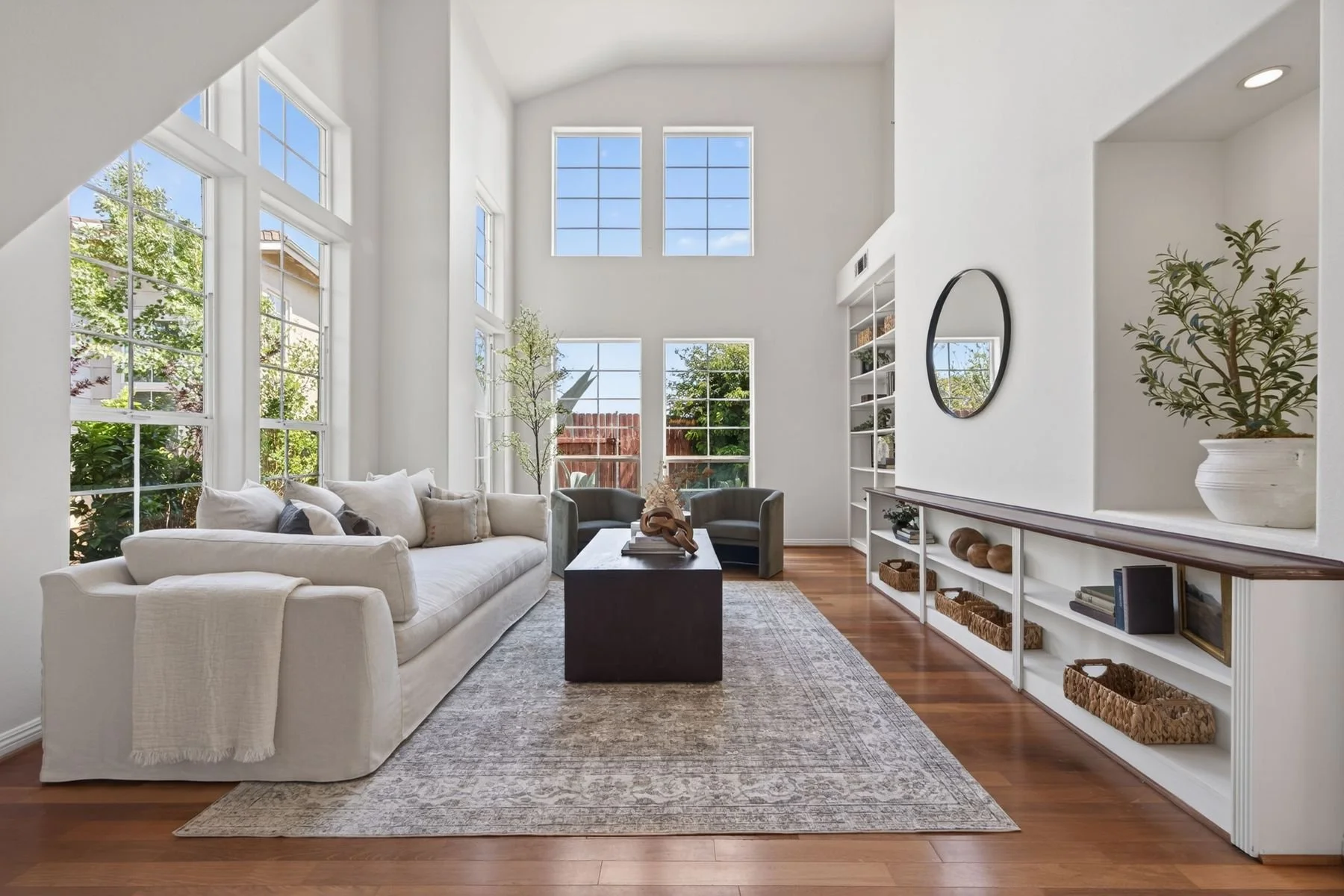 Bright living room with large windows, white walls, and high ceiling. Features a white sofa with beige and gray pillows, two gray armchairs, a black coffee table with decorative items, built-in white bookshelf with baskets and decor, a round mirror, 