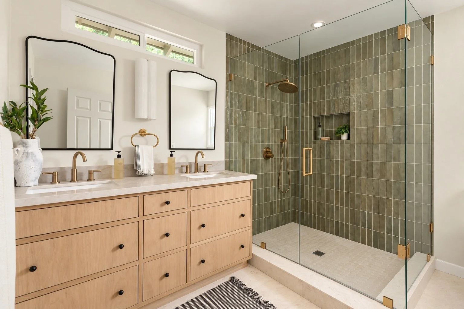 Modern bathroom with a double vanity with light wood cabinets, white countertop, gold fixtures, and two large rectangular mirrors. A glass-enclosed shower with green tiles, gold fixtures, and built-in shelf. A striped rug on a light-colored floor, an