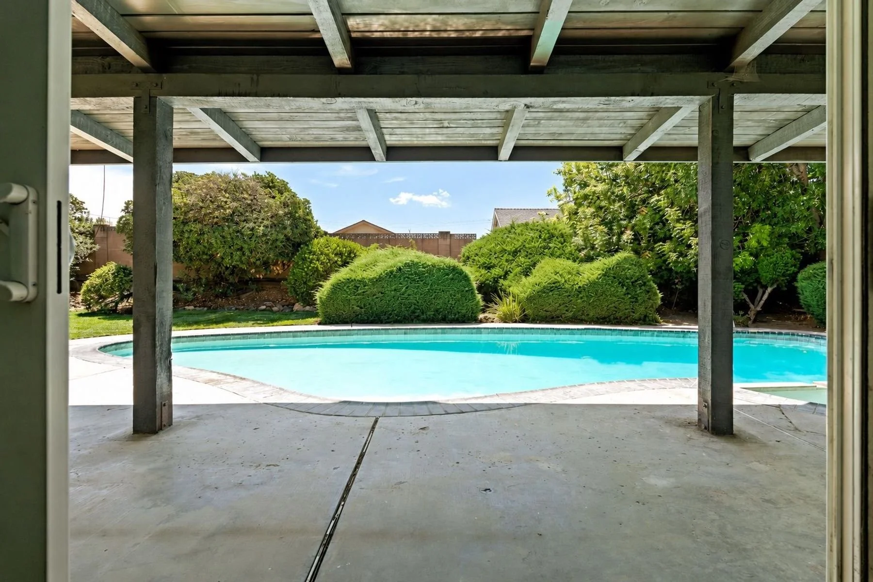 View of a backyard with a swimming pool, surrounded by bushes and trees, seen from underneath a covered patio.