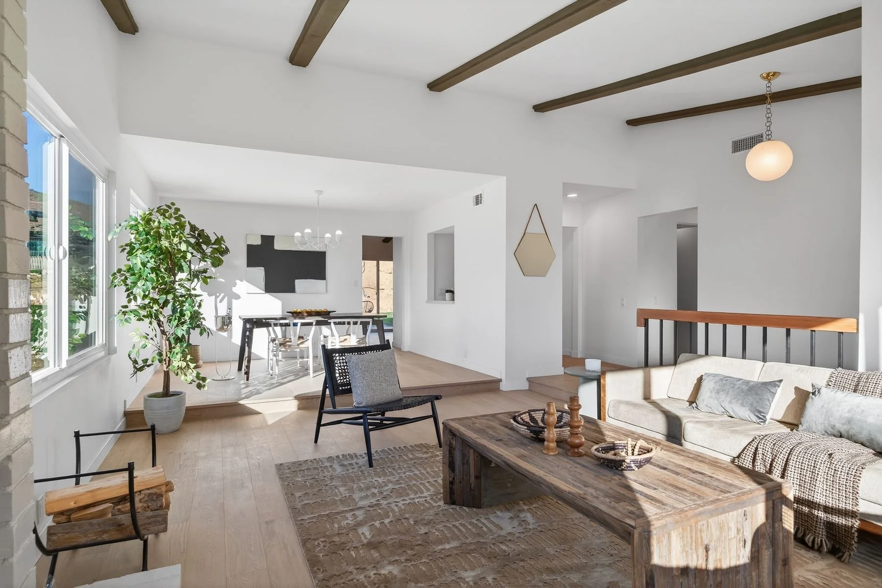 Living room with large window, plants, wooden flooring, sofa, coffee table, and dining area in background with a chandelier.