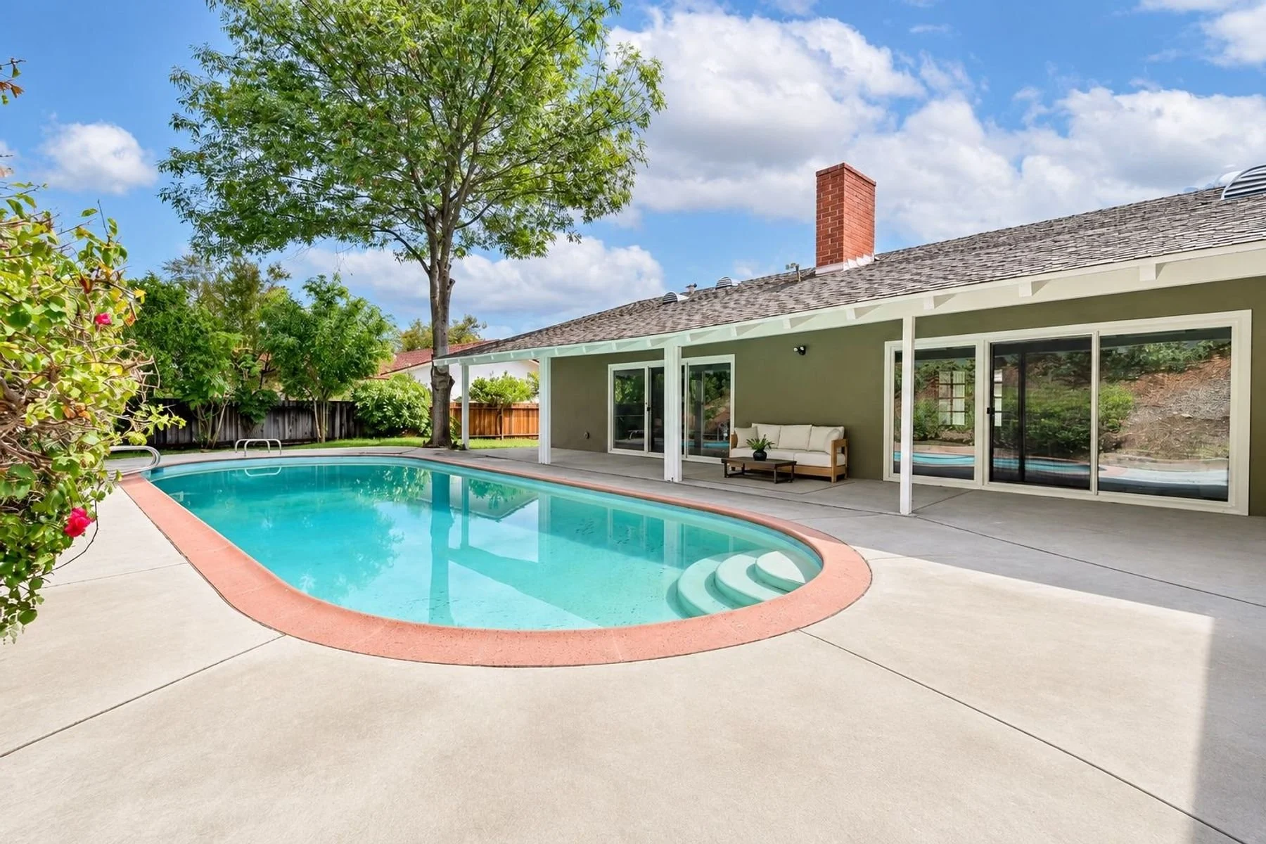 Backyard with a rectangular swimming pool, patio area with outdoor seating, large tree, wooden fence, house with sliding glass doors, and a partly cloudy sky.