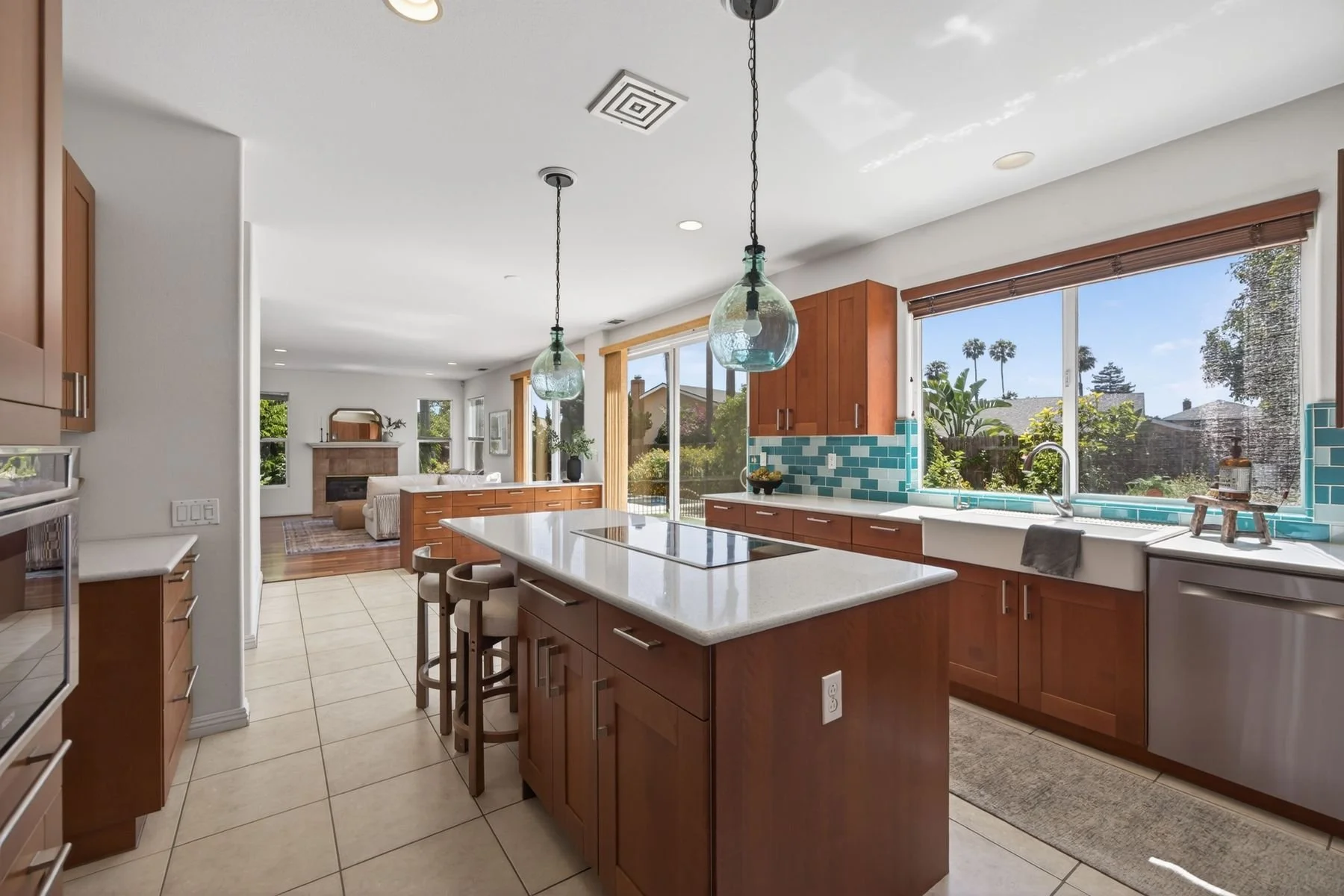 Modern kitchen with wooden cabinets, white countertops, turquoise tile backsplash, large window with a view of trees, island with seating, and pendant lights hanging above.