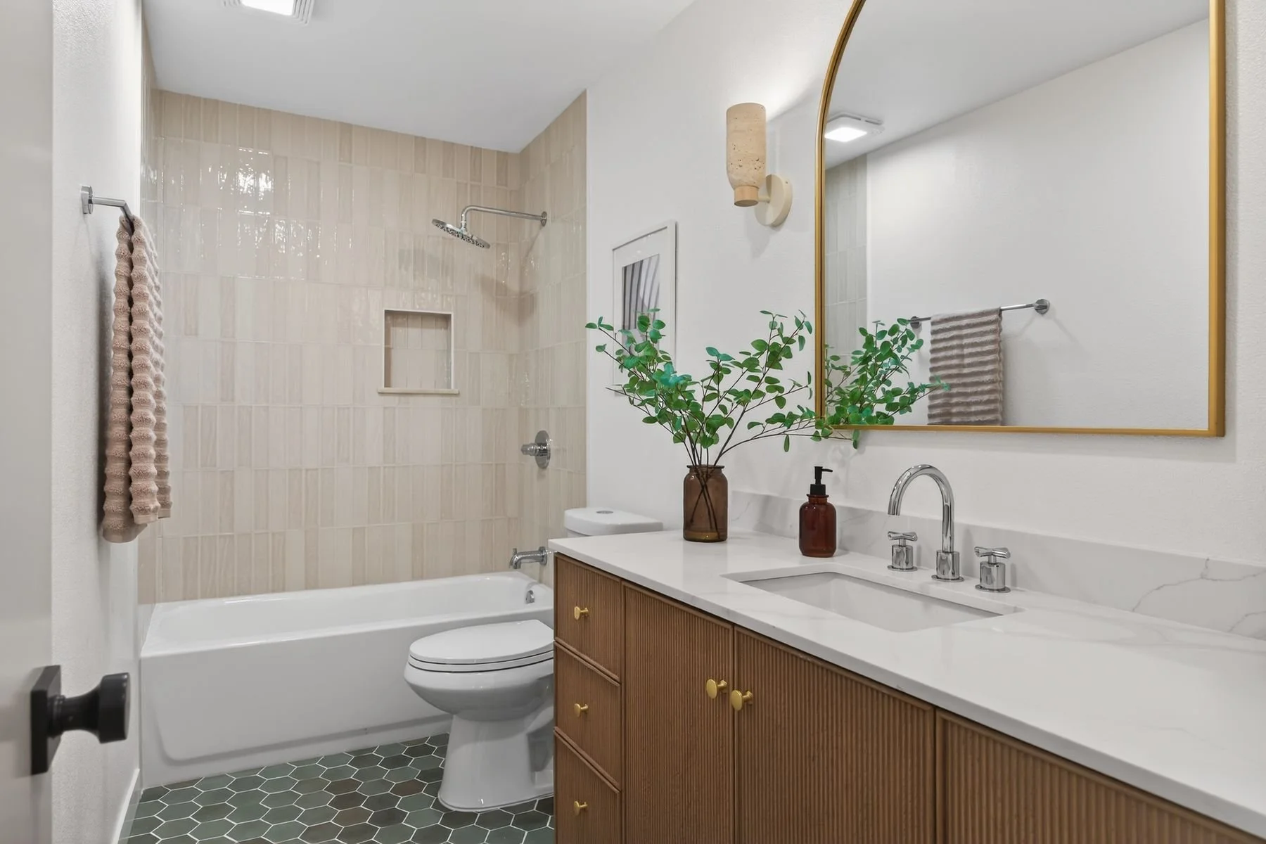 A bathroom with a bathtub and shower on the left, a toilet in the center, and a wooden vanity with a white countertop on the right. The vanity has a sink, a silver faucet, and brown and amber bottles. A large mirror with a gold frame hangs above the 