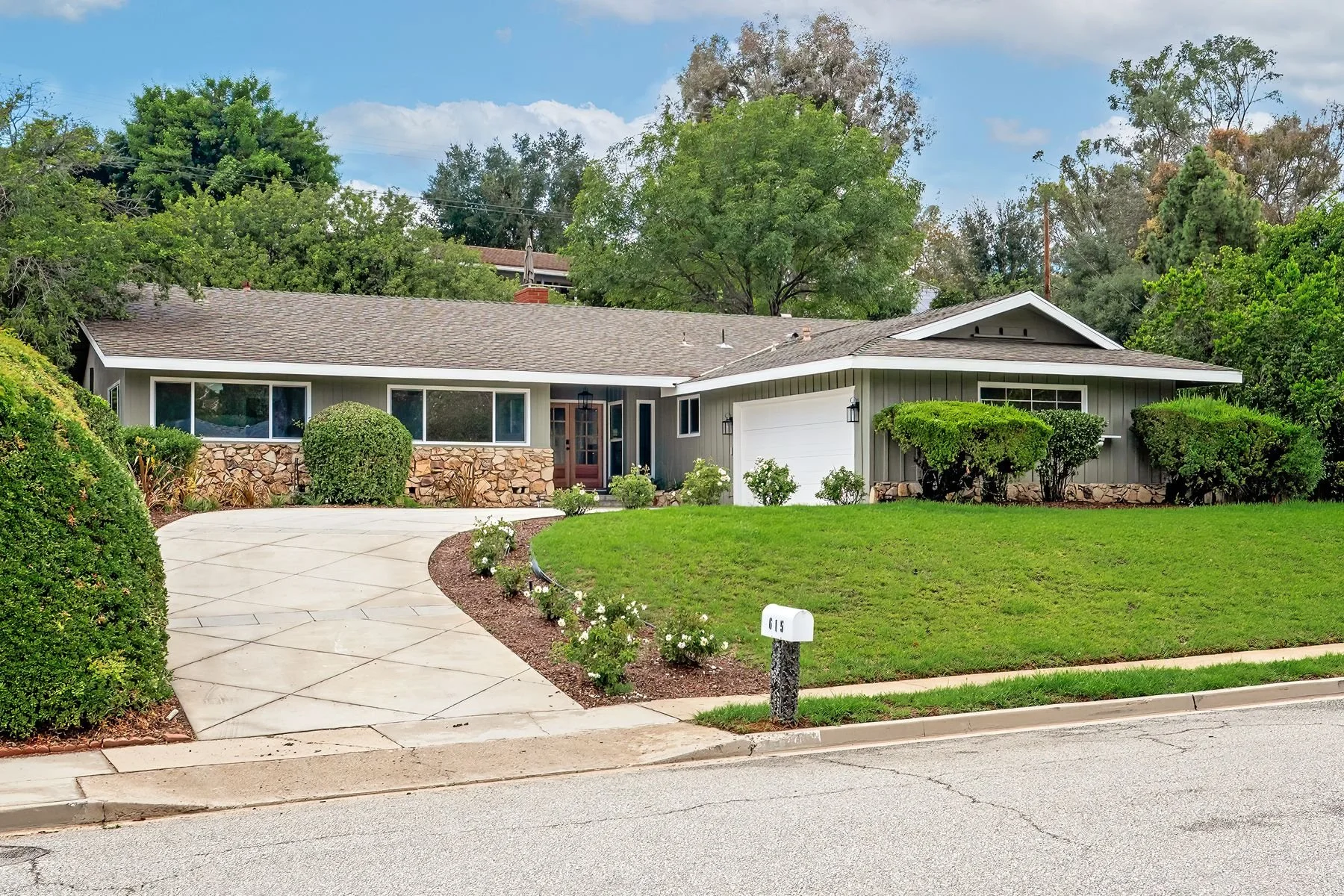 front view of a single-story house with a stone and panel exterior, a white garage door, a decorative front door, surrounded by a well-maintained green lawn and landscaped bushes, with a curved concrete driveway and a street in the foreground, and ta